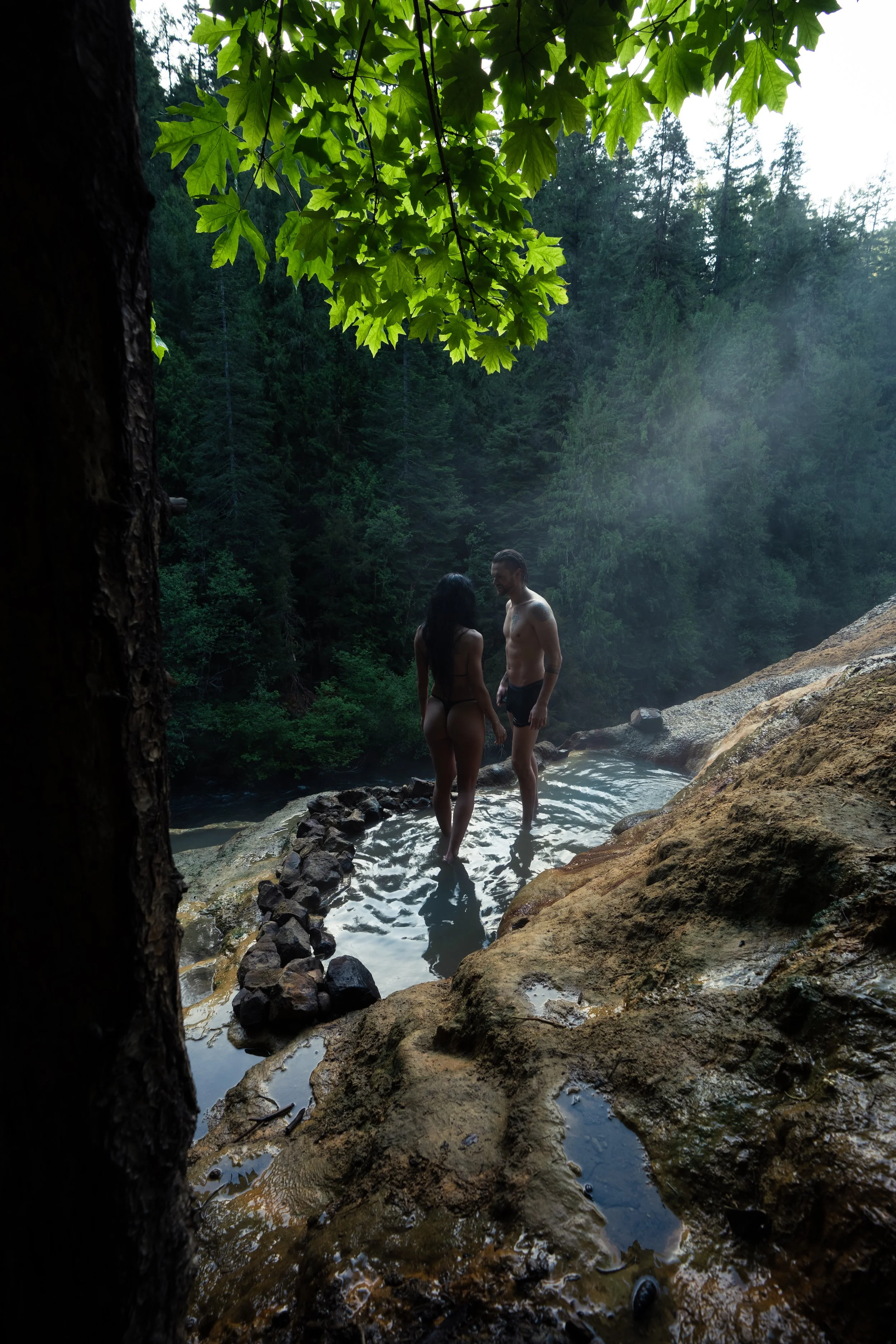 A man and woman standing in a natural hot spring or waterfall surrounded by lush greenery and trees.