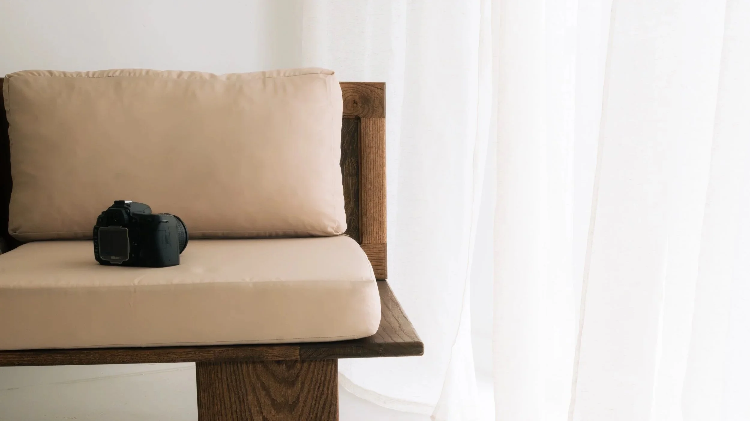 A black digital camera resting on a beige cushion on a wooden bench, with a white sheer curtain and sunlight in the background.