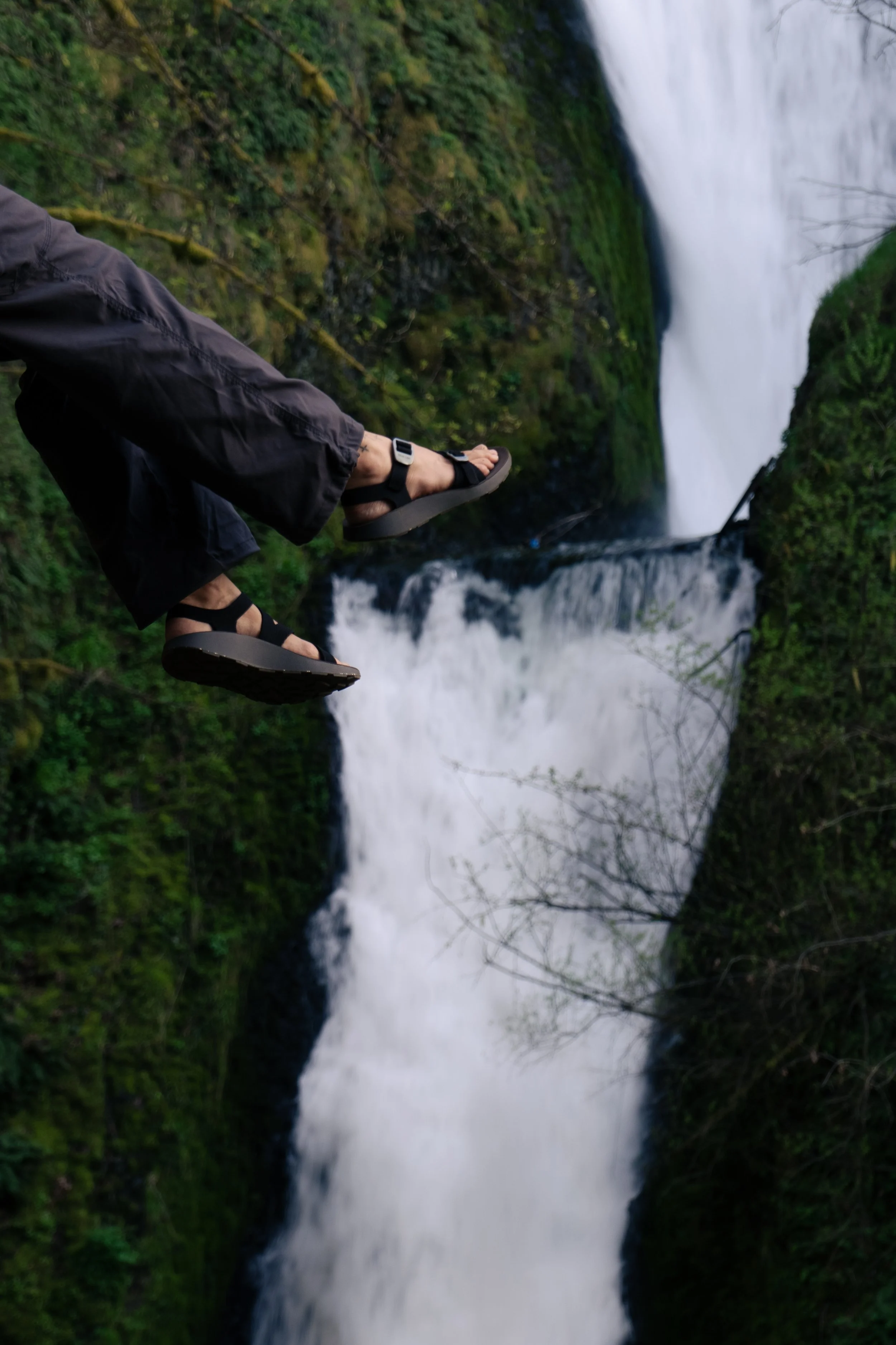 Person wearing black pants and black sandals standing on a ledge with a waterfall in the background.