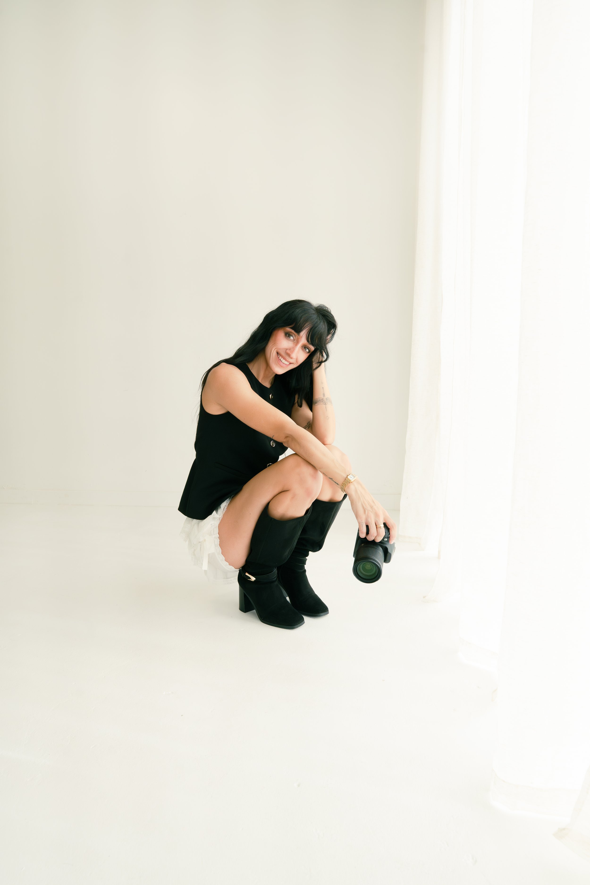 Woman with black hair in black top and white shorts squatting on white floor, holding a camera, smiling, near white curtains.