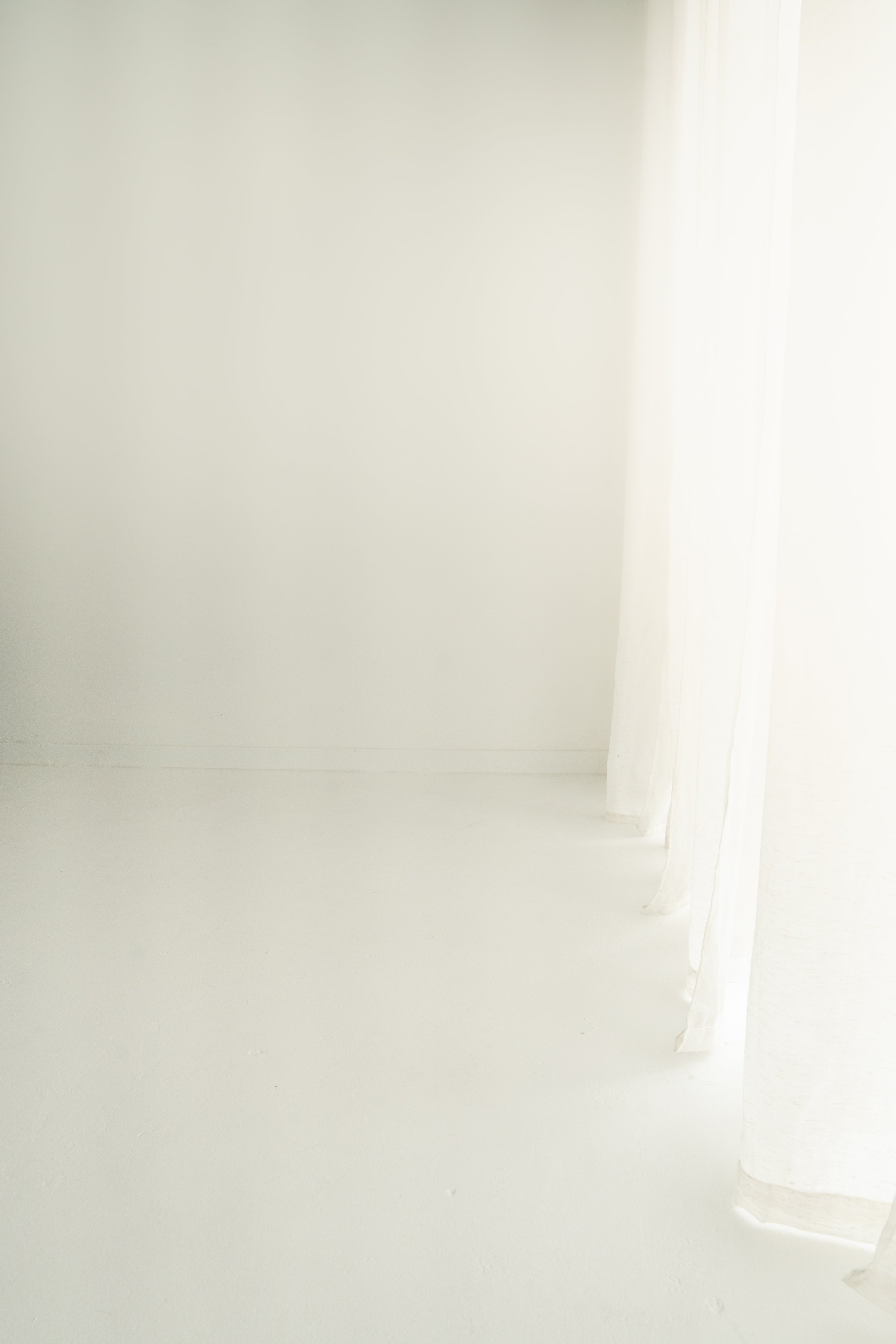 Empty white room with sheer white curtains hanging by a window.