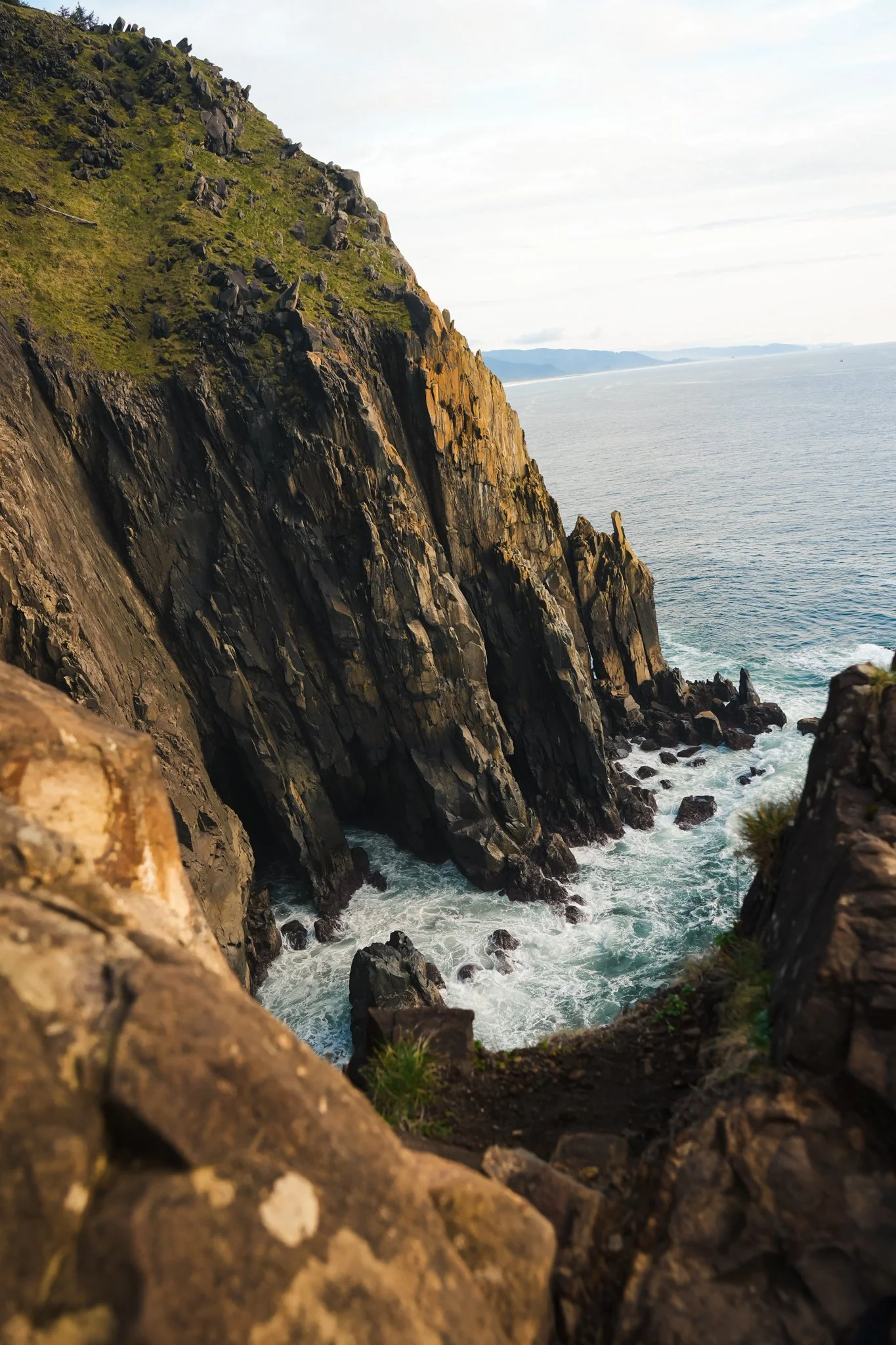 Coastal cliffs with rugged, dark rocks and green patches, ocean waves crashing at the base, and a hazy horizon in the background.