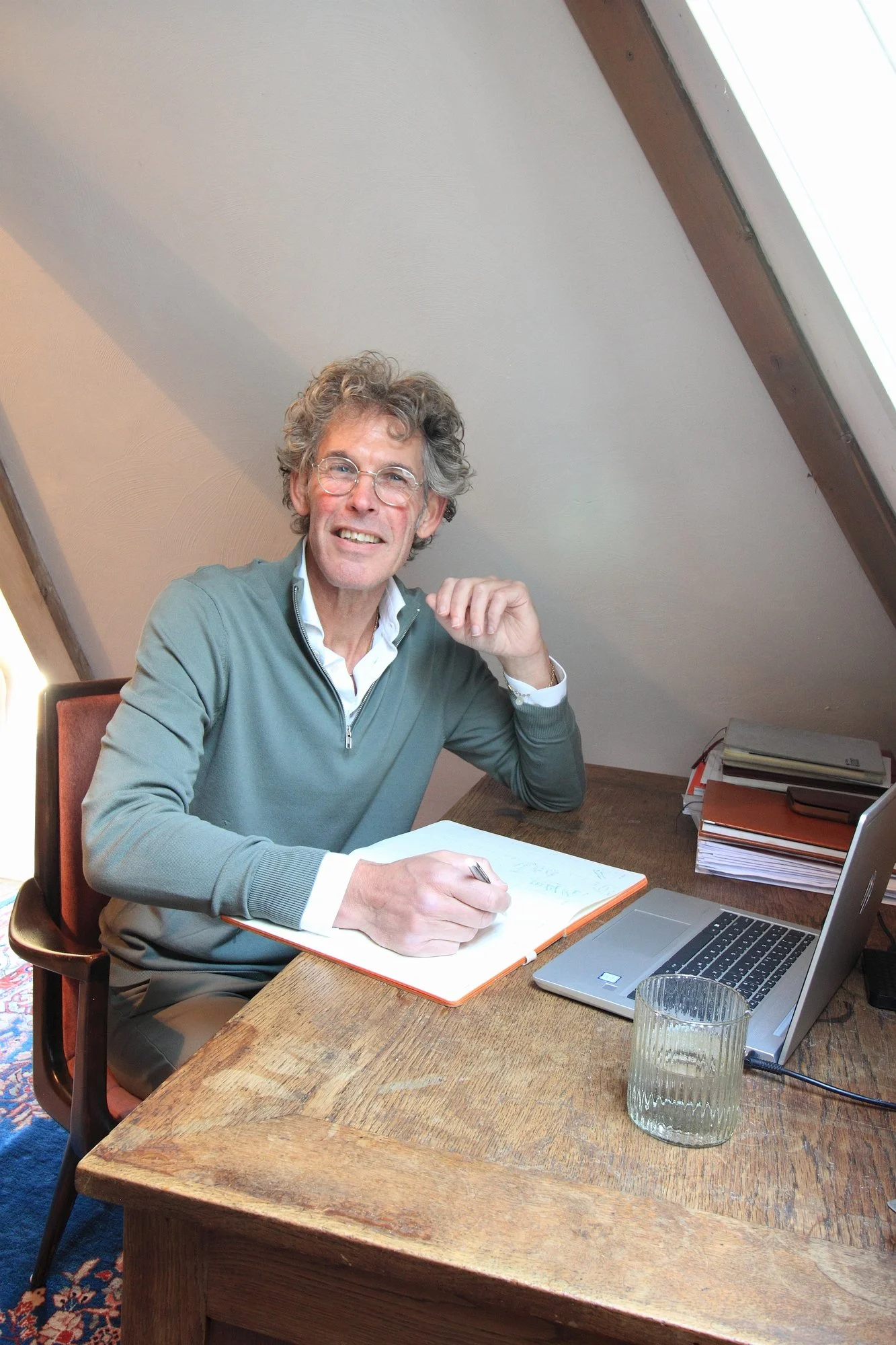 Man met glazen bril schrijft in een notitieboekje aan een houten bureau met een laptop, een glas water en stapels paperassen.