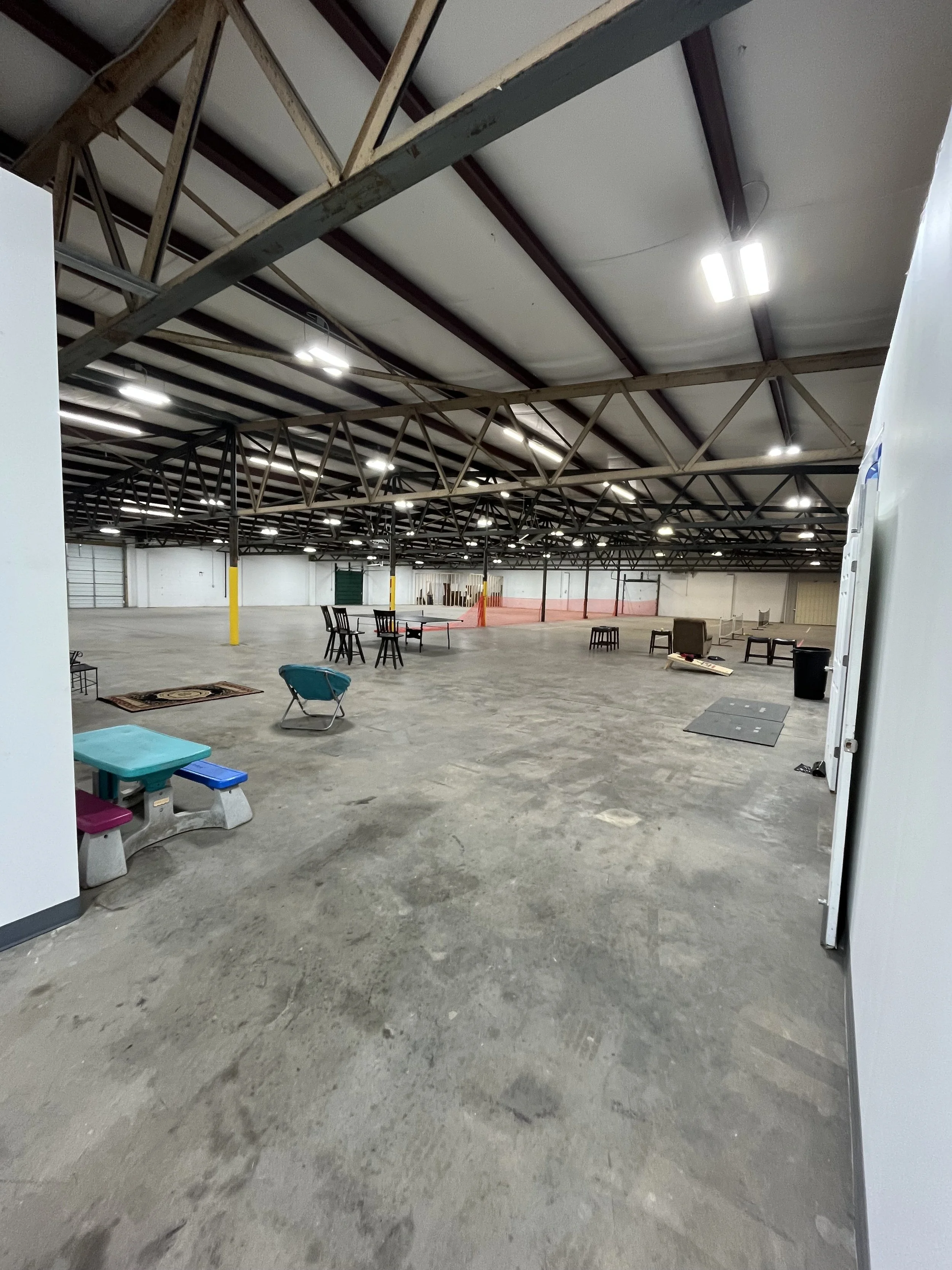 Empty warehouse with scattered furniture, including chairs, small tables, and a picnic bench, under a metal roof with bright overhead lighting.