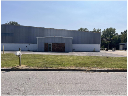 Small commercial building with a metal facade, brick accents, and a side entrance, situated in a grassy area with a parking lot and a street in front.