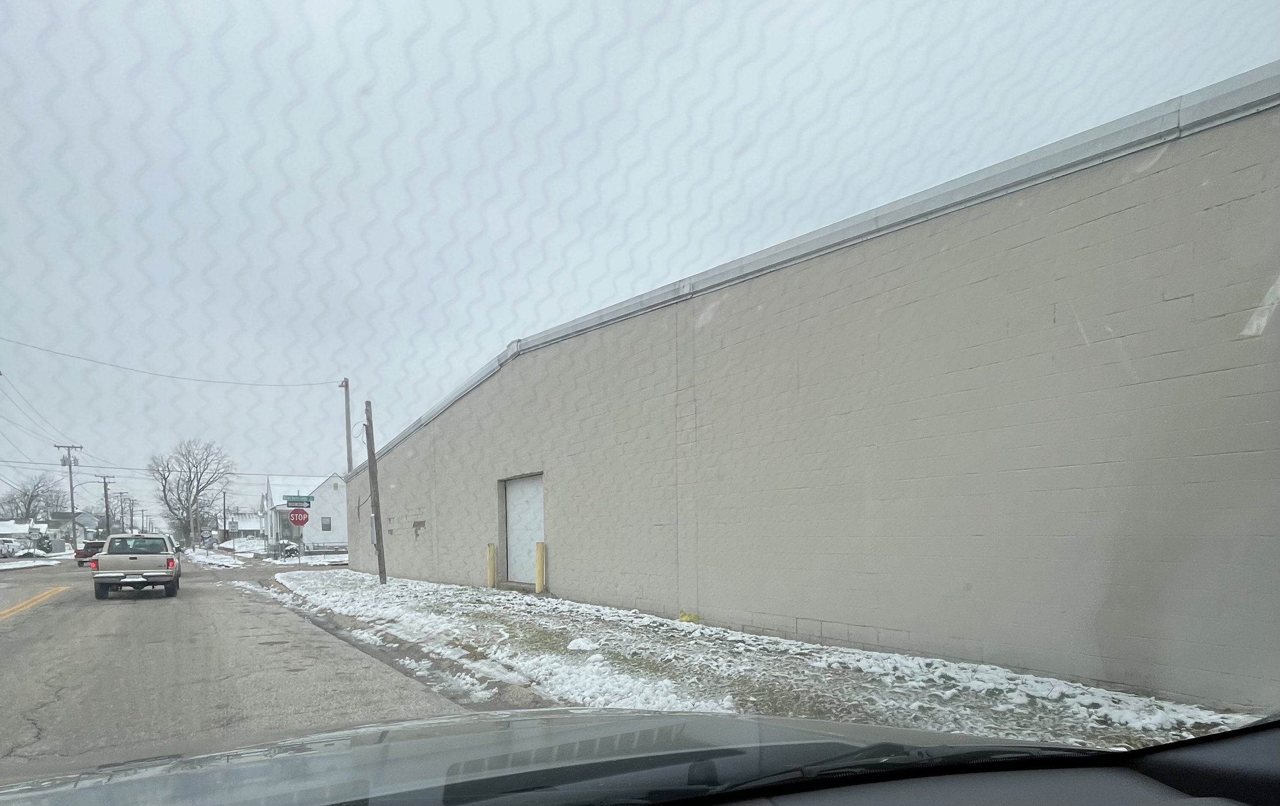 View from inside a vehicle looking through a windshield on a snowy day, showing a street with cars, a stop sign, utility poles, and a large beige industrial building with a garage door.