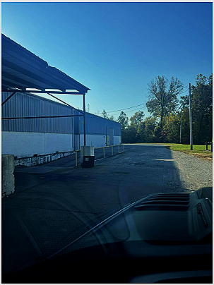 An empty parking lot near a white building with a metal roof, with trees and blue sky in the background.