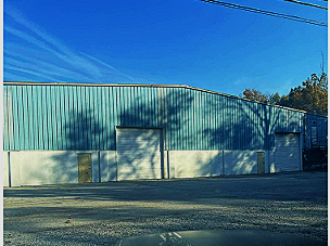A large industrial warehouse with a green metal exterior and a white concrete lower section, two closed garage doors, under a clear blue sky.