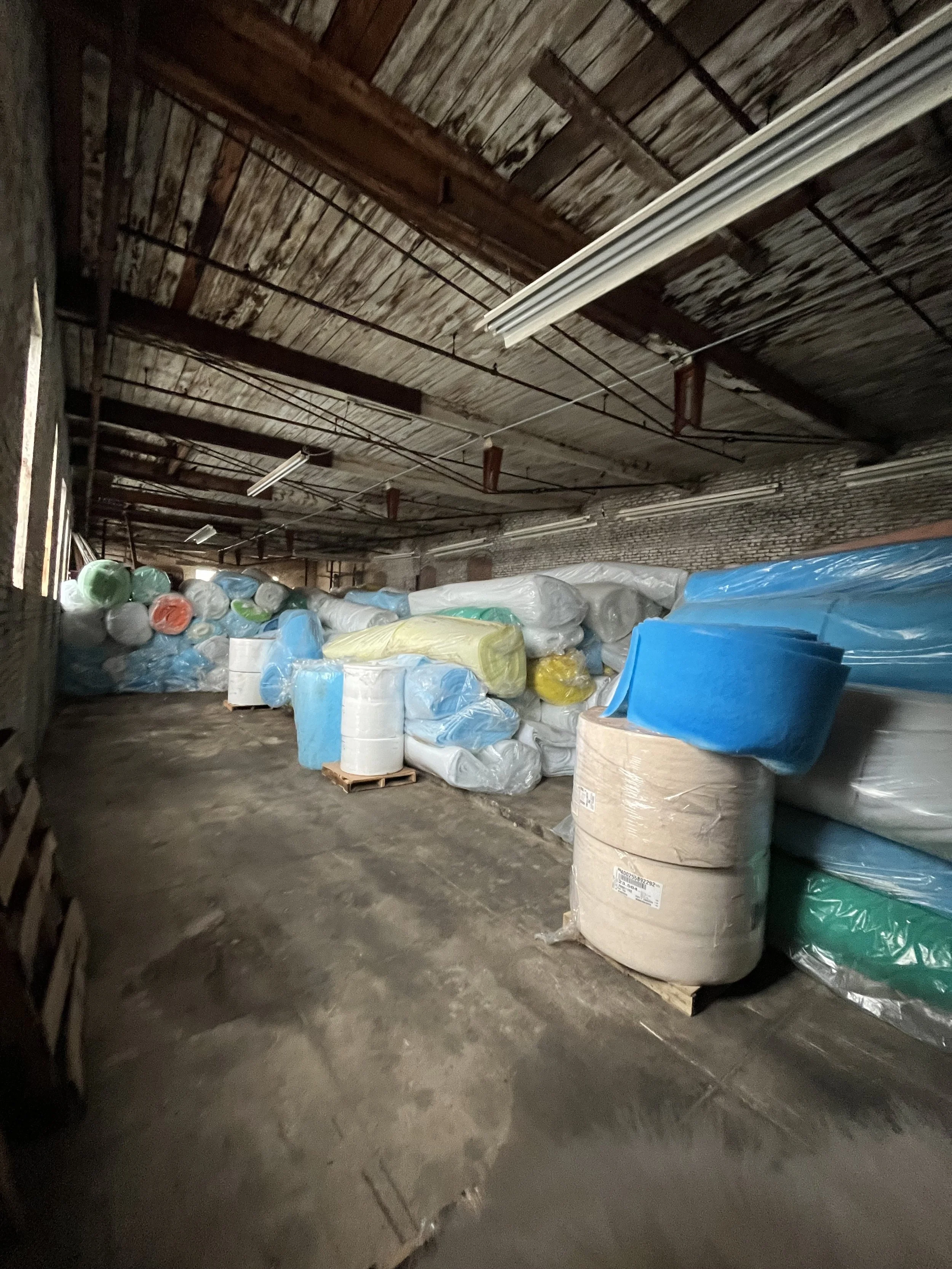 Storage room with plastic-wrapped rolls of fabric or insulation stacked along the wall, on a concrete floor and ceiling with exposed wooden beams.