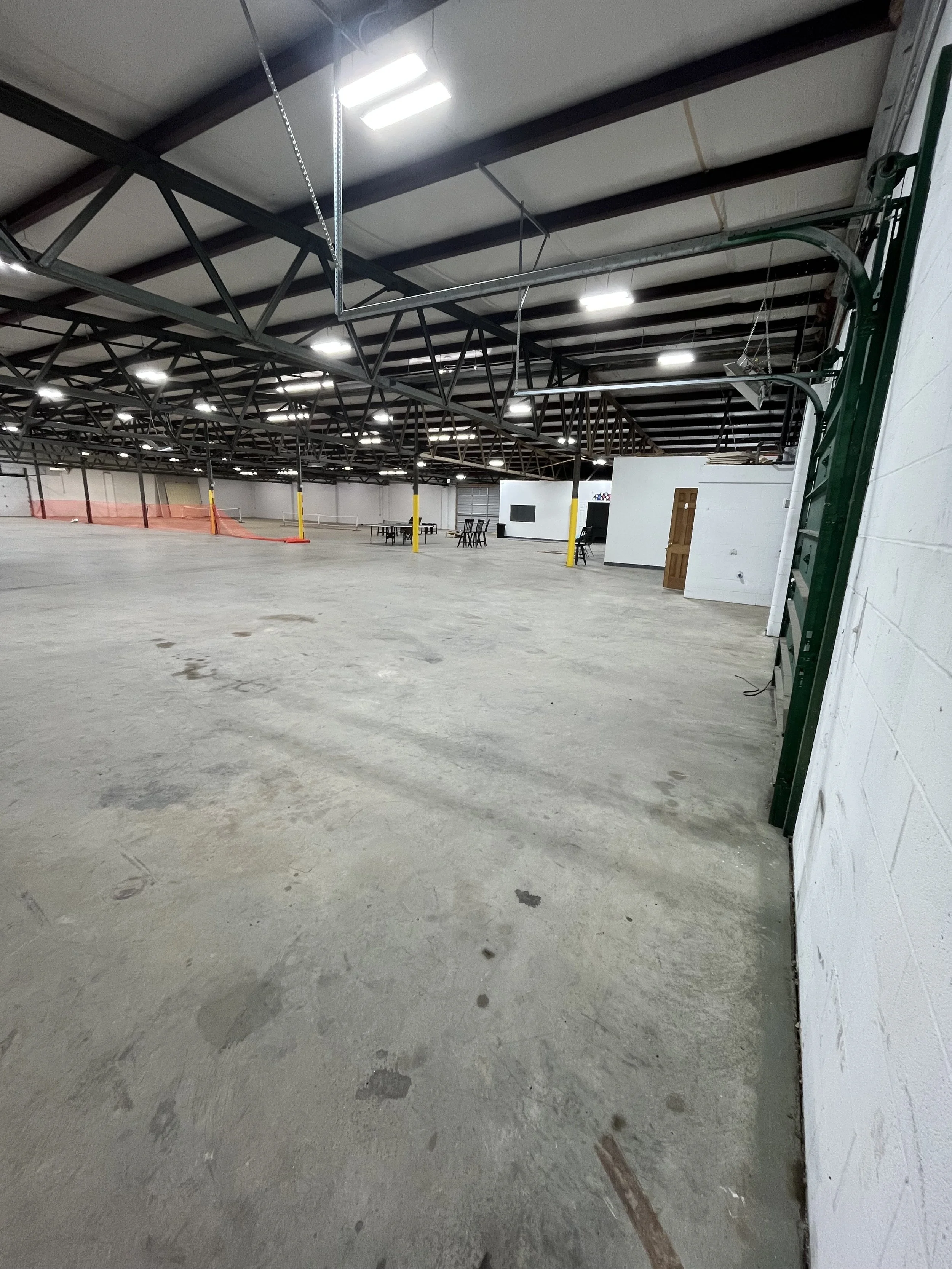 Empty warehouse space with concrete floor, metal ceiling, overhead lights, some furniture, and a partially visible green door frame.