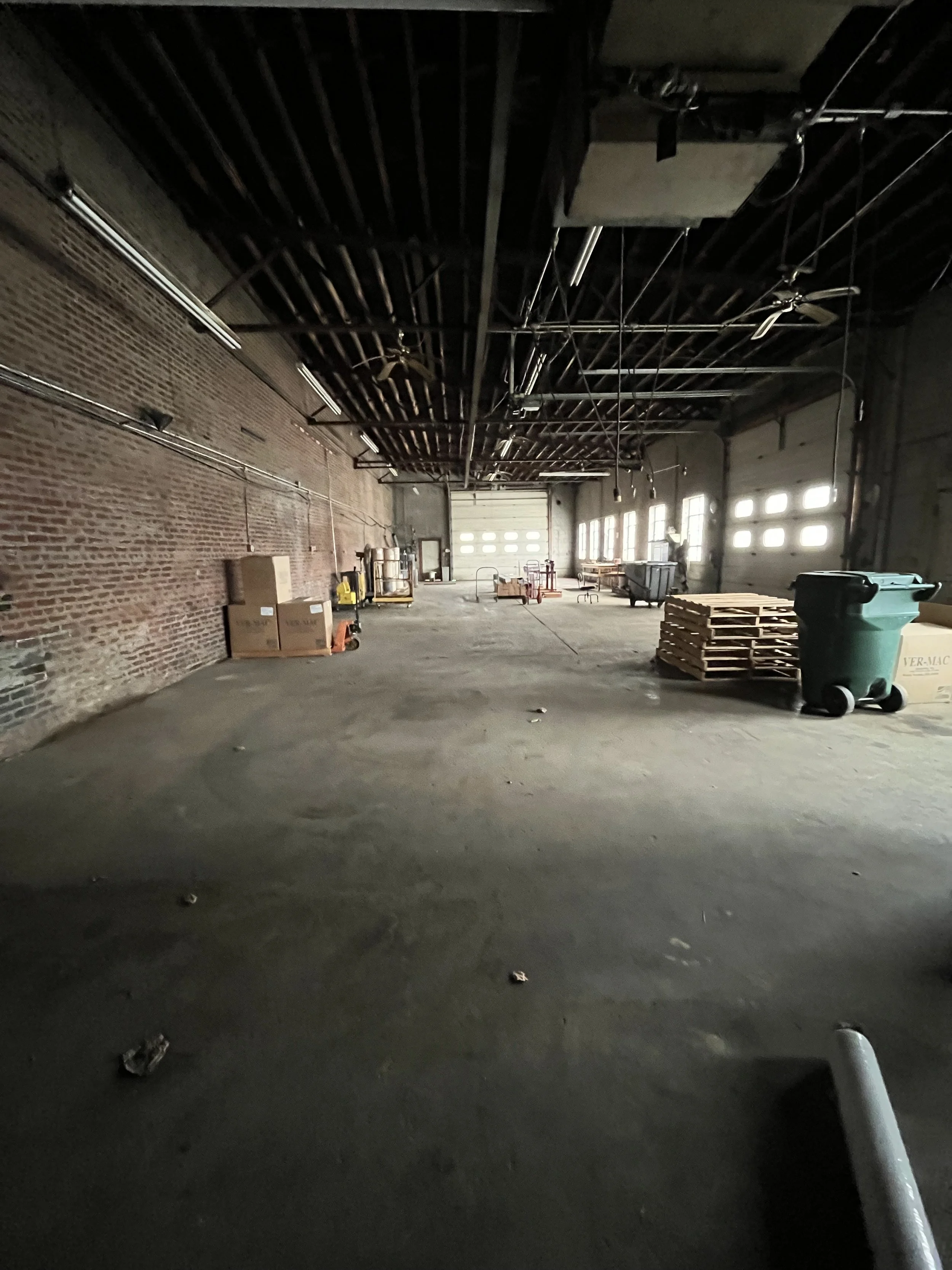 Empty industrial warehouse with brick walls, high ceiling, and large garage door. Scattered pallets, containers, and equipment on the concrete floor.