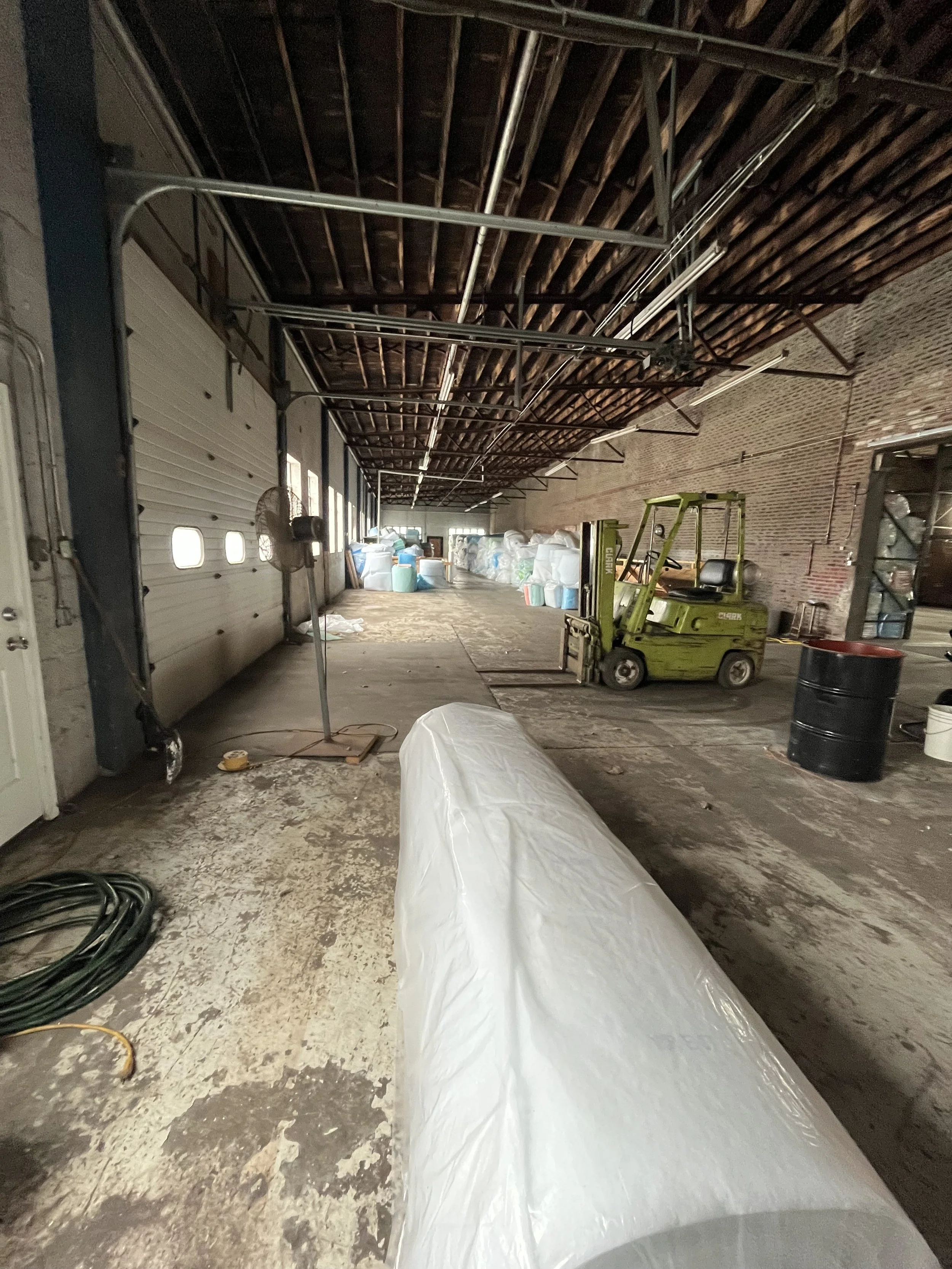 Interior of a warehouse with a concrete floor, empty space extending into the background, and various equipment including a green forklift, large blue and white barrels, and rolled plastic or fabric in the foreground. Exposed rafters and electrical c