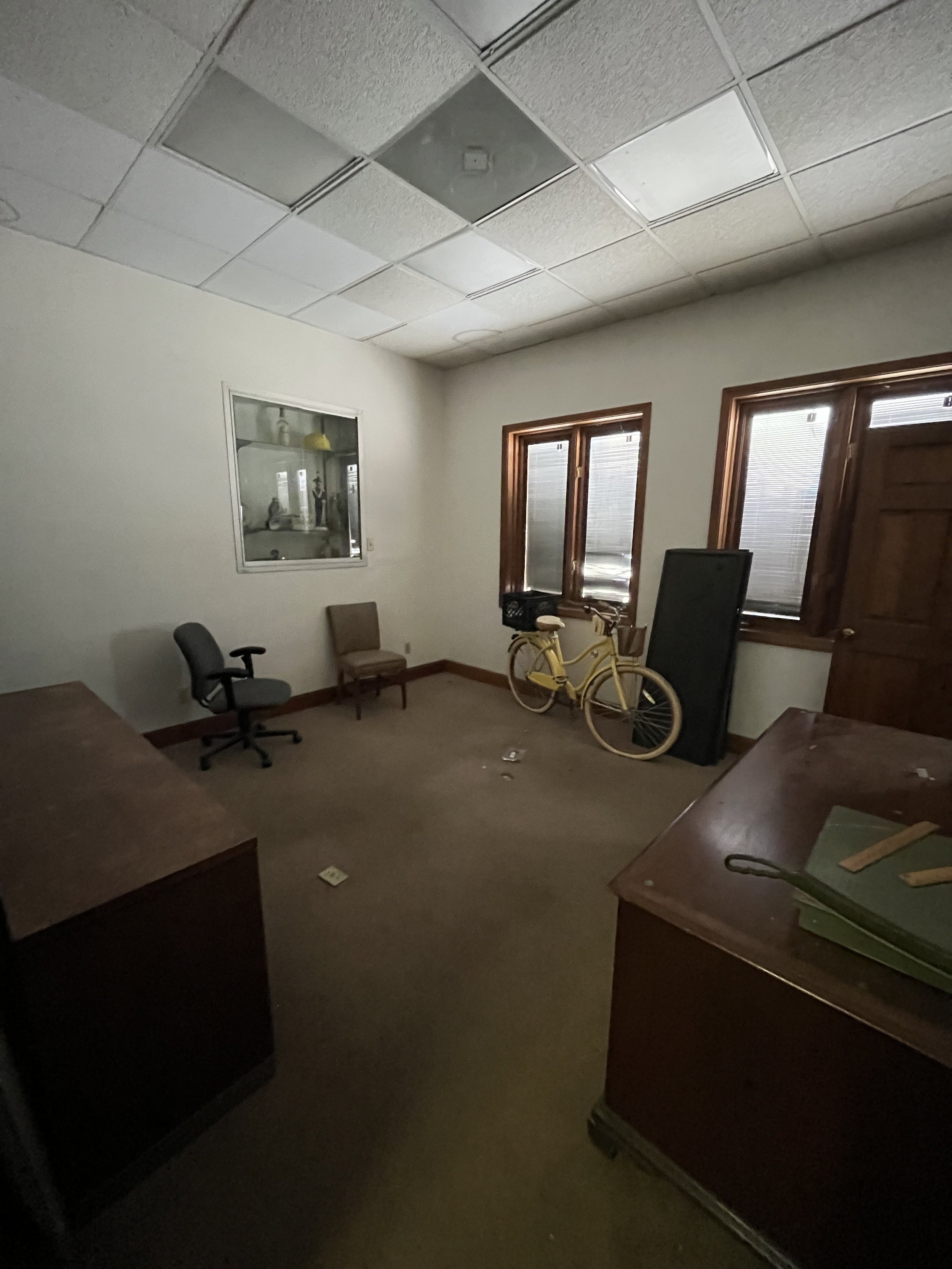 Empty office room with two windows, a yellow bicycle, two chairs, a black screen, desks, and scattered items on the carpeted floor.
