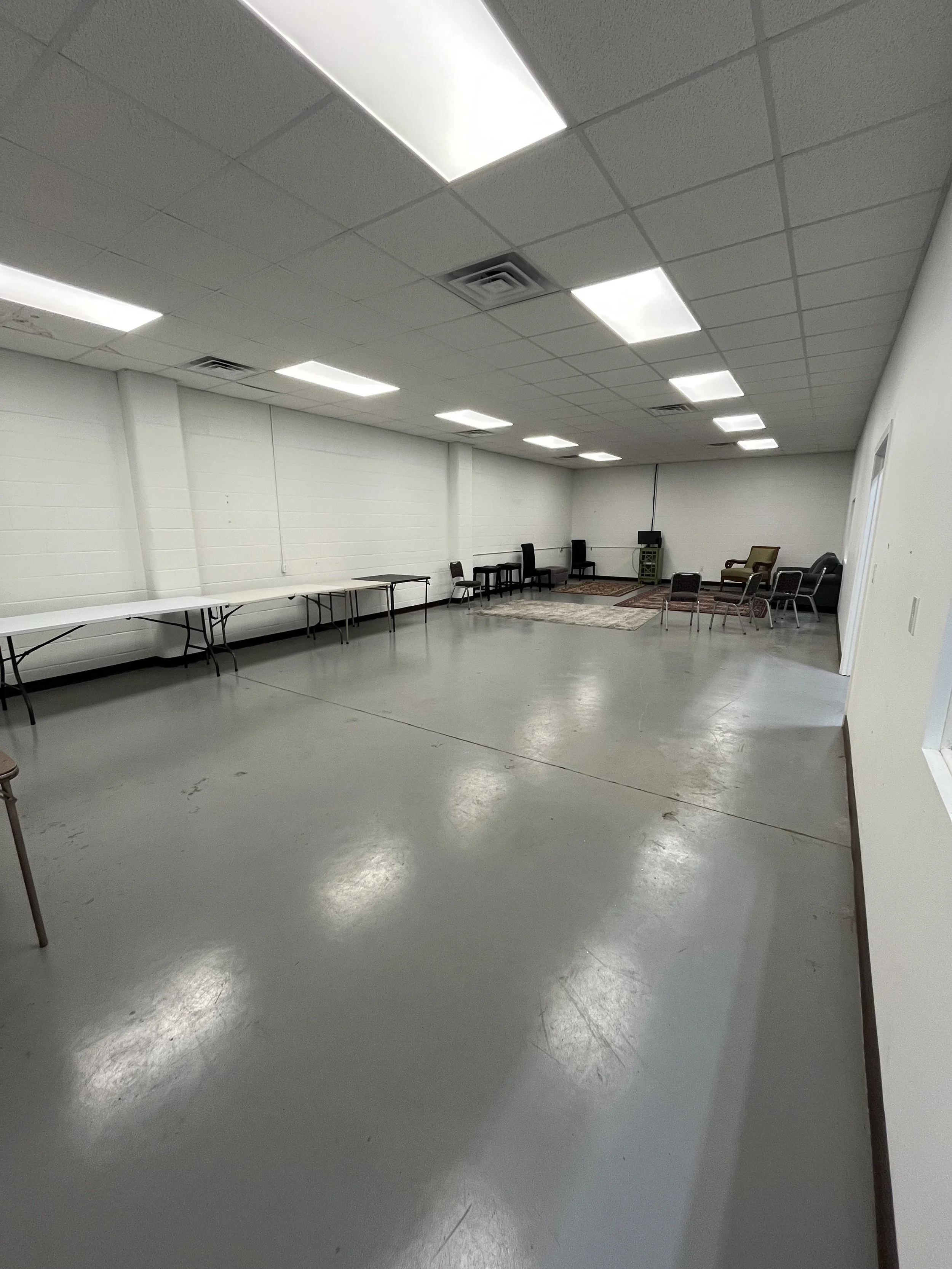 Empty multipurpose room with folding tables and chairs, white walls, tiled ceiling, and fluorescent lighting.
