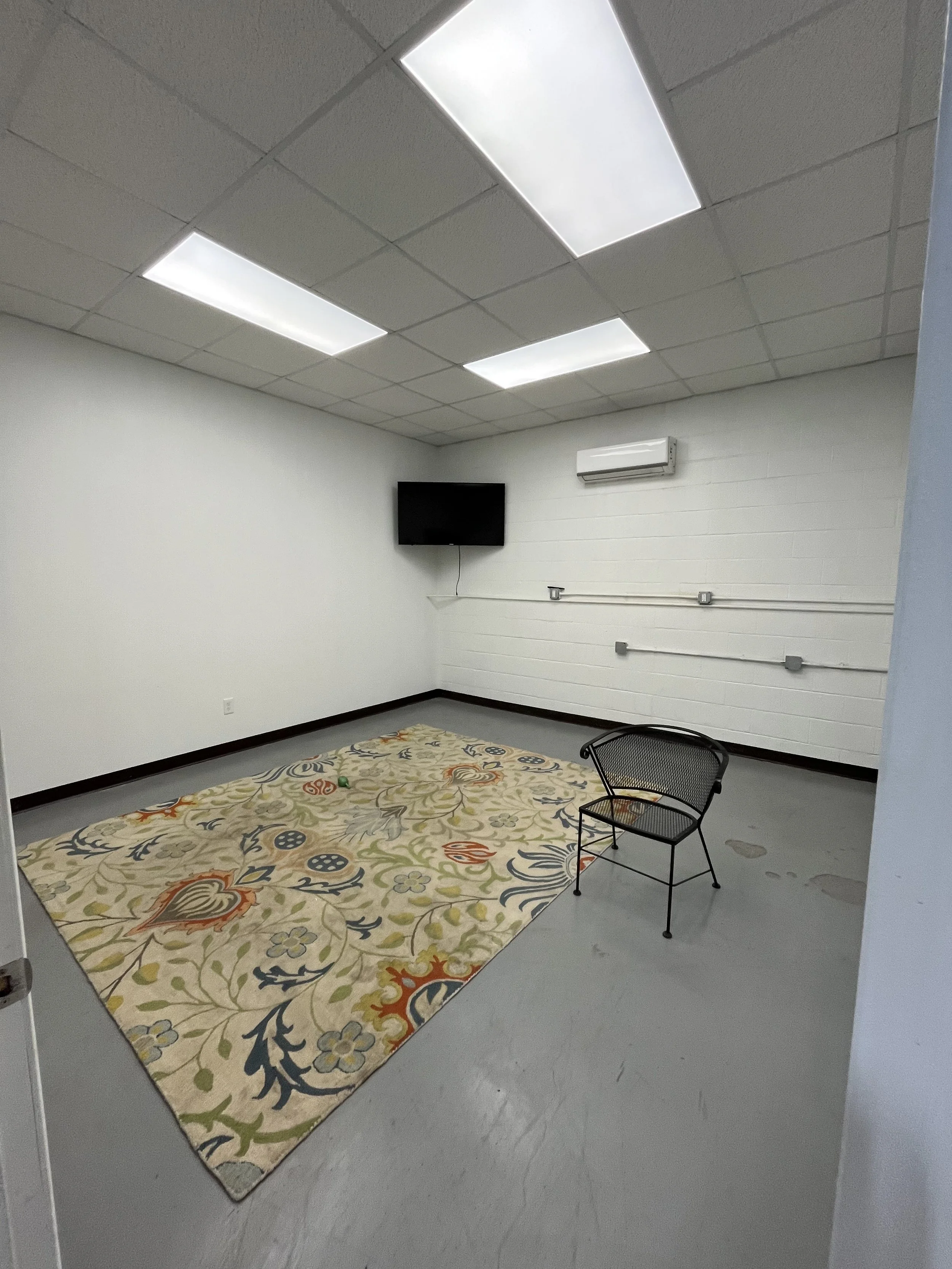 Empty room with a colorful floral rug, black chair, wall-mounted television, air conditioning unit, and fluorescent ceiling lights.