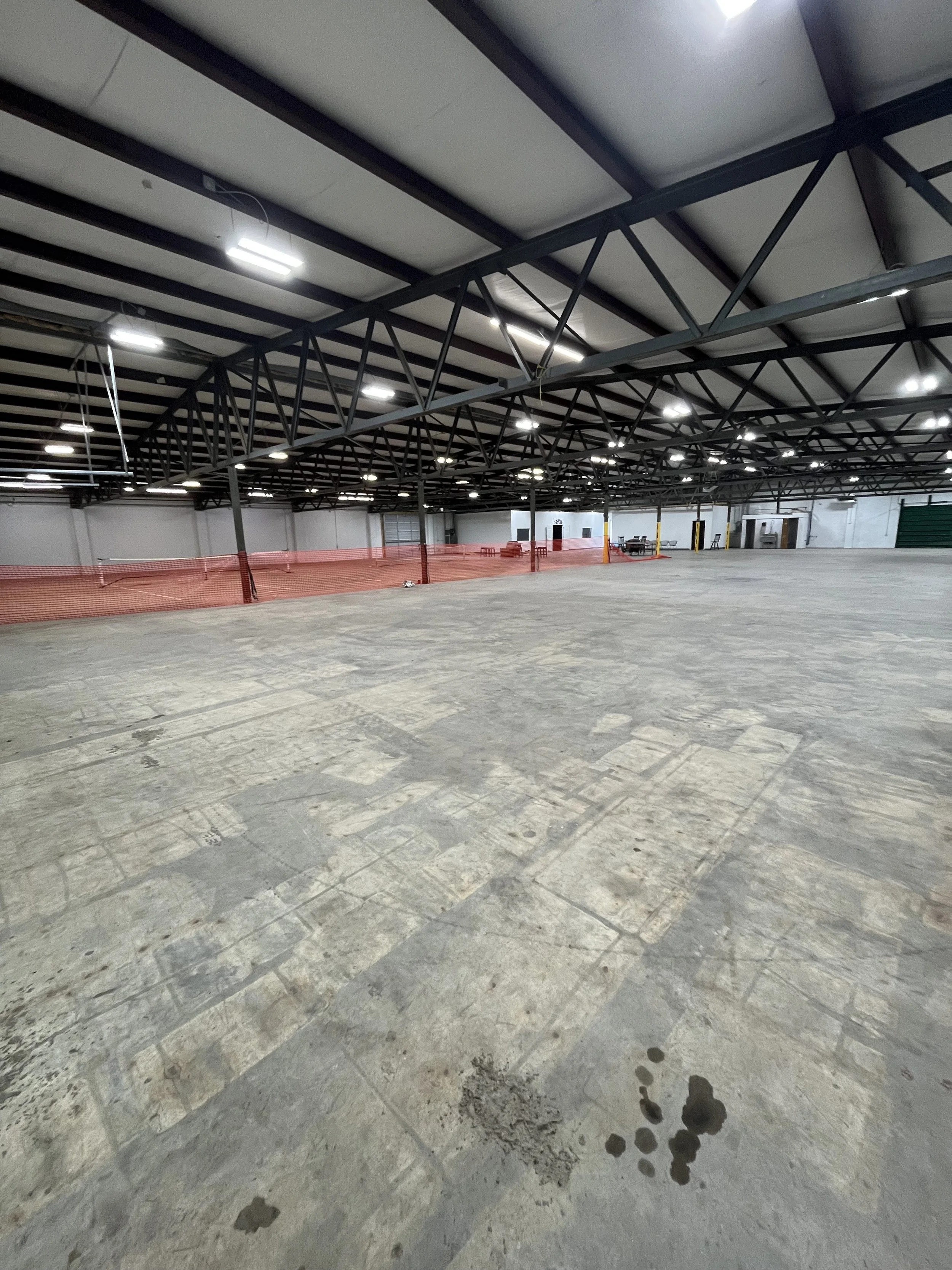 Empty indoor warehouse with concrete floor, exposed steel beams on ceiling, bright overhead lighting, and orange safety netting in the background.