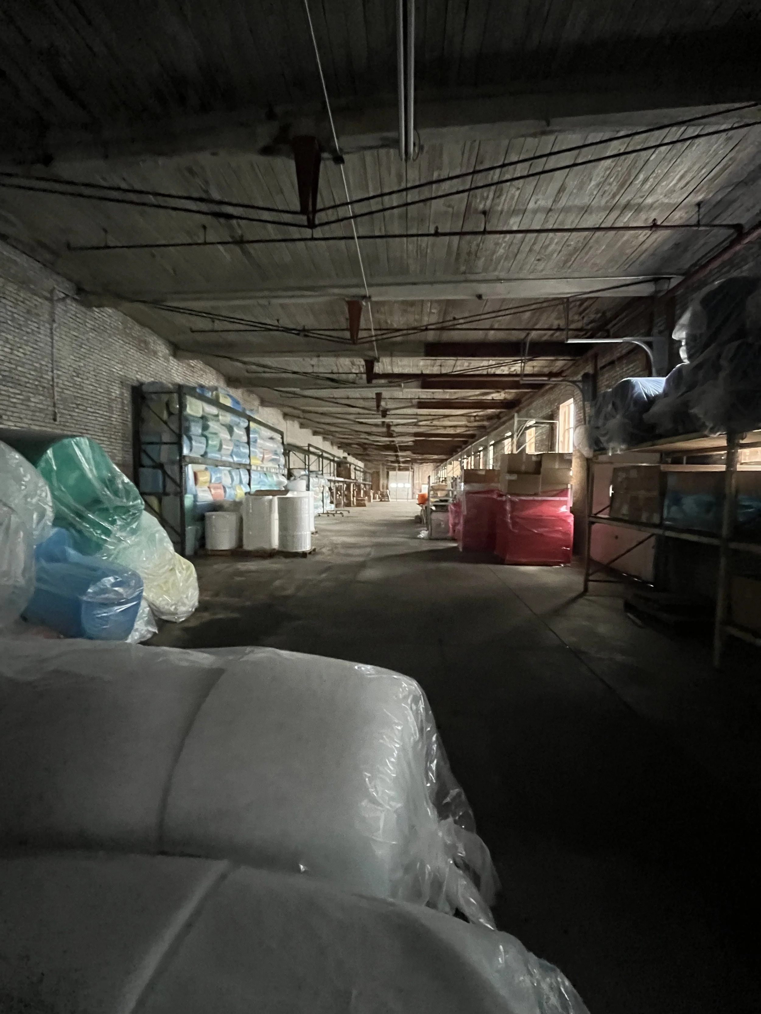 View of an industrial or storage space with shelves containing various supplies, wrapped pallets, large plastic-wrapped items, and a long aisle with windows letting in natural light.
