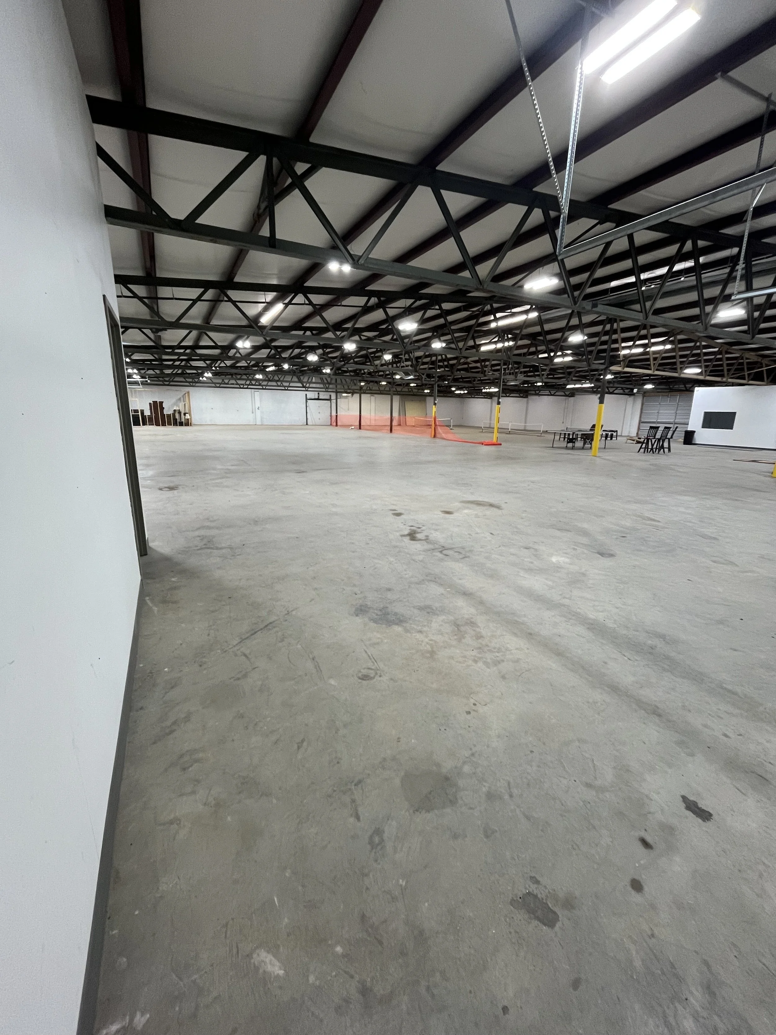 Empty warehouse or large indoor space with unfinished concrete floor, exposed ceiling beams, and some scattered tables and chairs in the background.