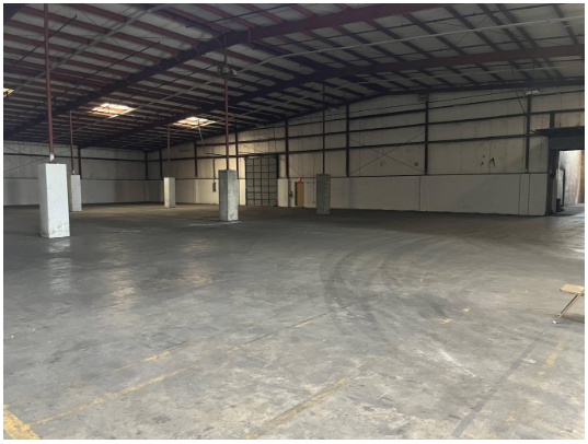 Empty warehouse with concrete floor and metal roof, supported by white pillars, with a few visible doors and no vehicles or people.