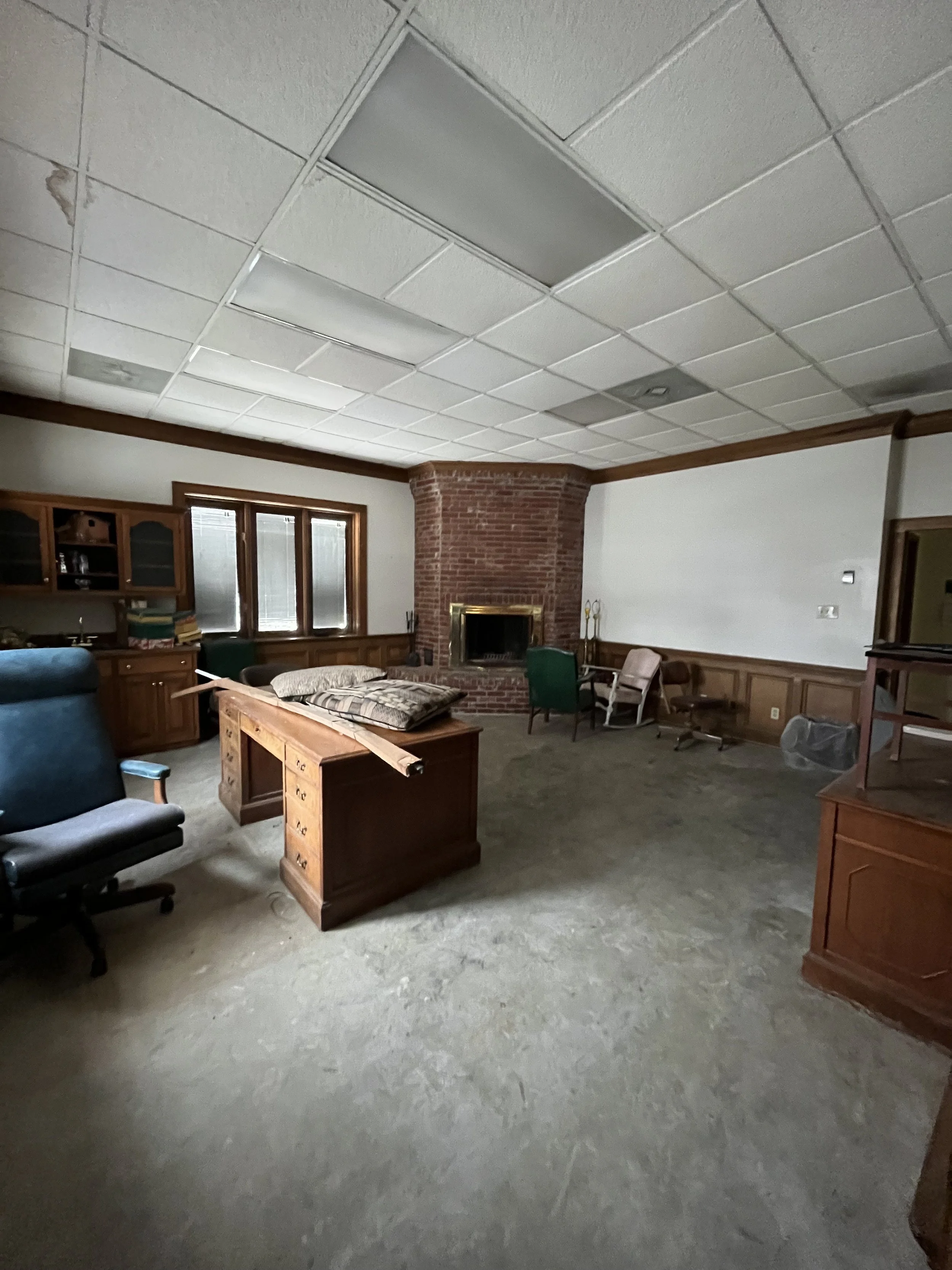 Office or living room with a brick fireplace, wooden furniture, office chair, and cluttered desk.