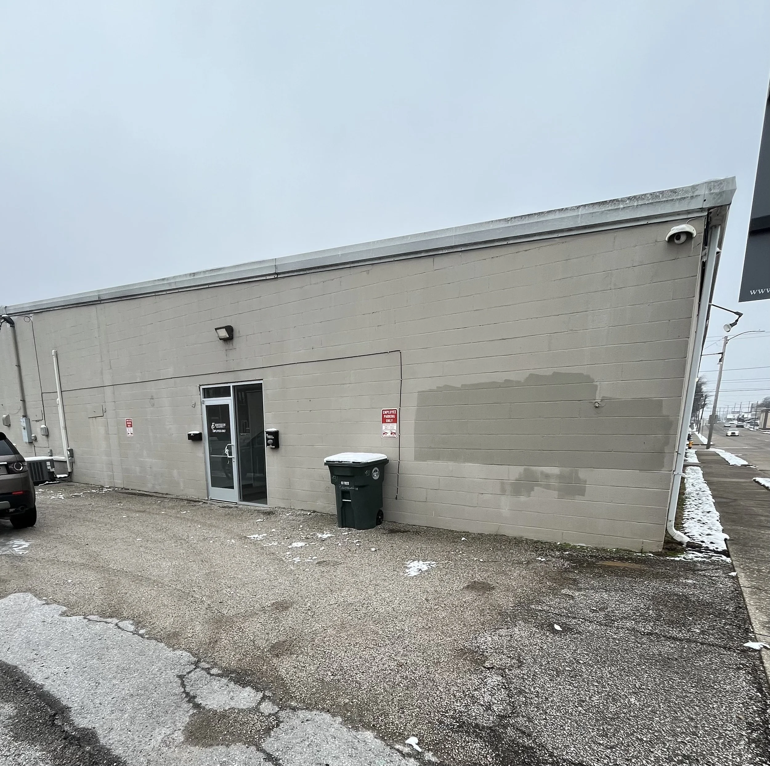 A beige commercial building with a single glass door, two black mailboxes, a green trash bin, and a small parking area with patches of snow and patches of asphalt. There are signs and security cameras on the building.