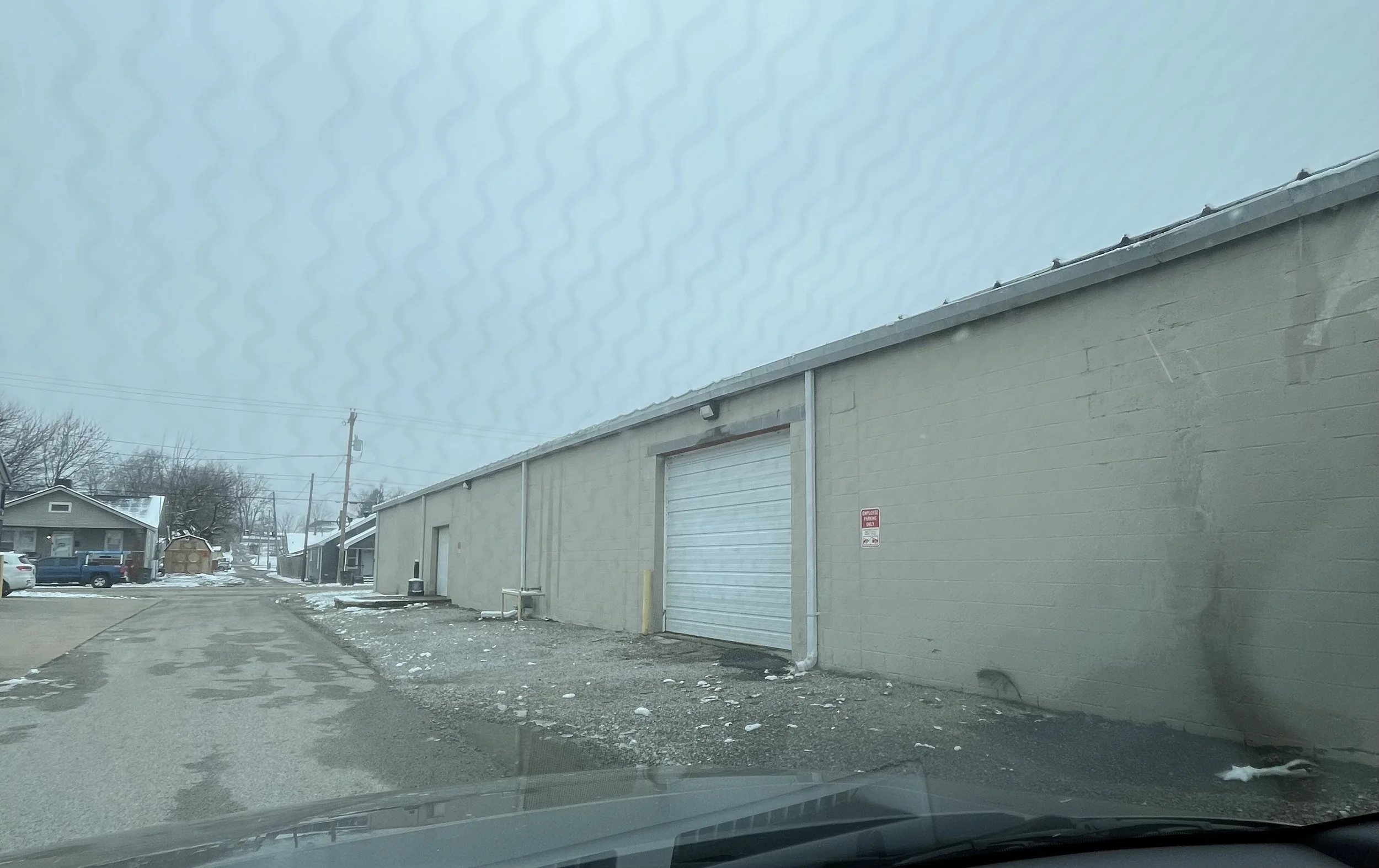 Photo of a gray industrial building with a roll-up door, located in a parking lot with snow and ice, with residential houses and trees in the background, taken on a cloudy winter day.