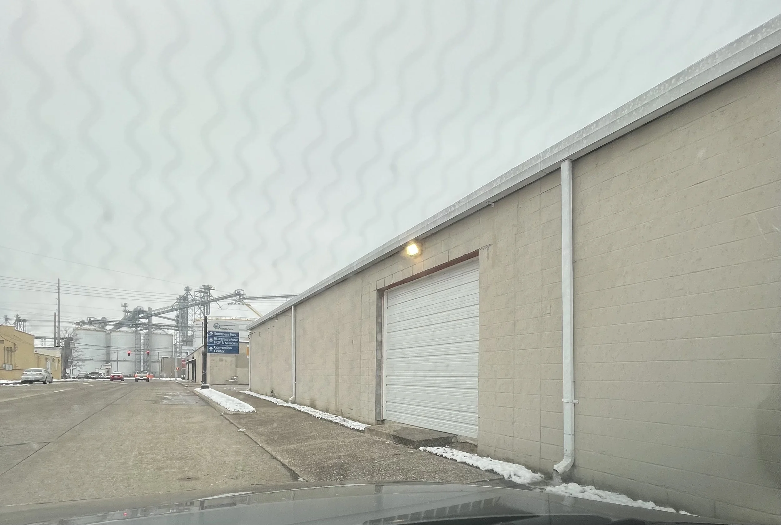 A large industrial building with a closed white roll-up door, a street light, and snow along the sidewalk, with a parking lot and industrial tanks and structures in the background on a cloudy day.
