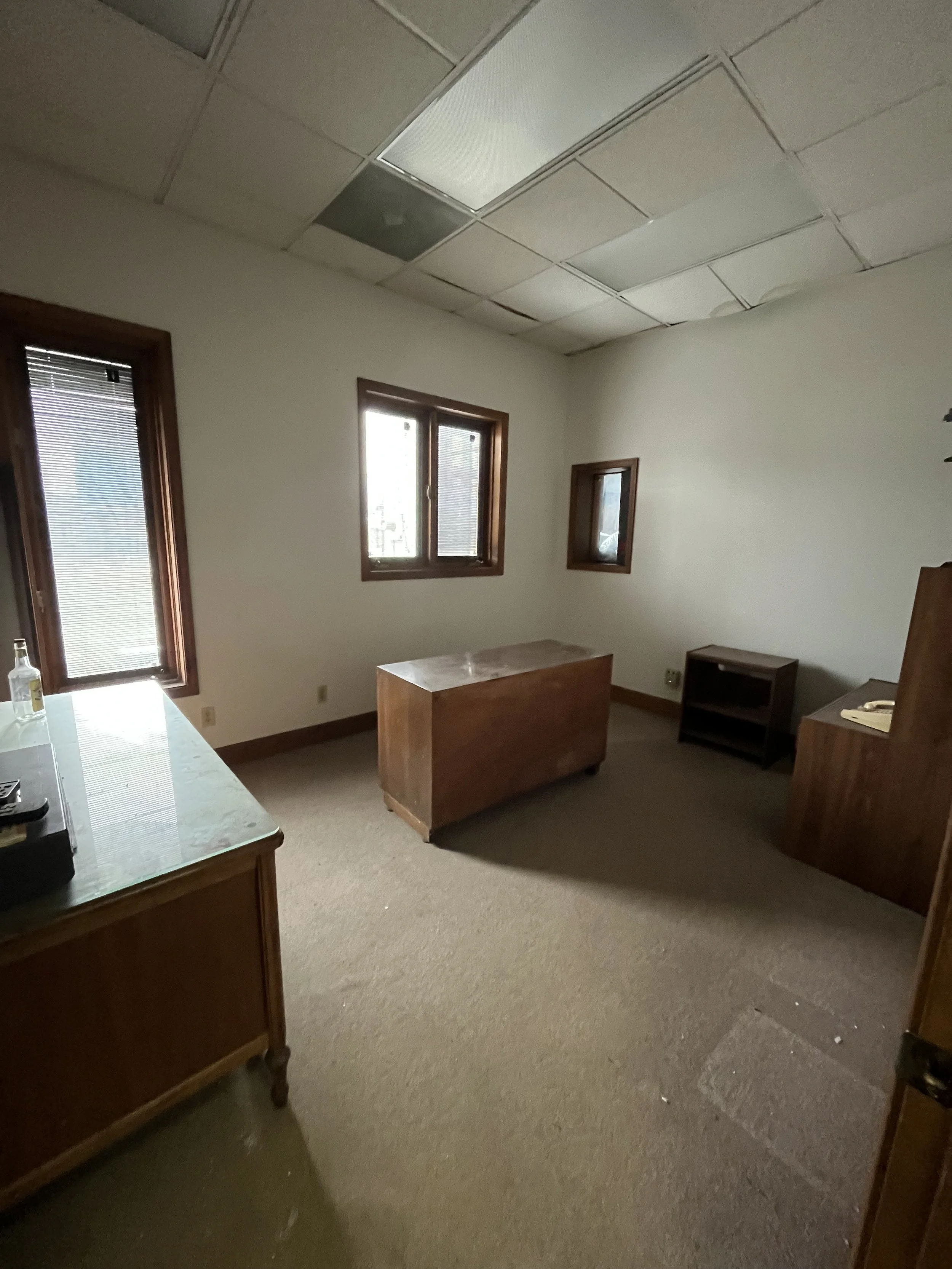 Empty office room with three windows, wooden furniture, and a ceiling with missing or damaged ceiling tiles.