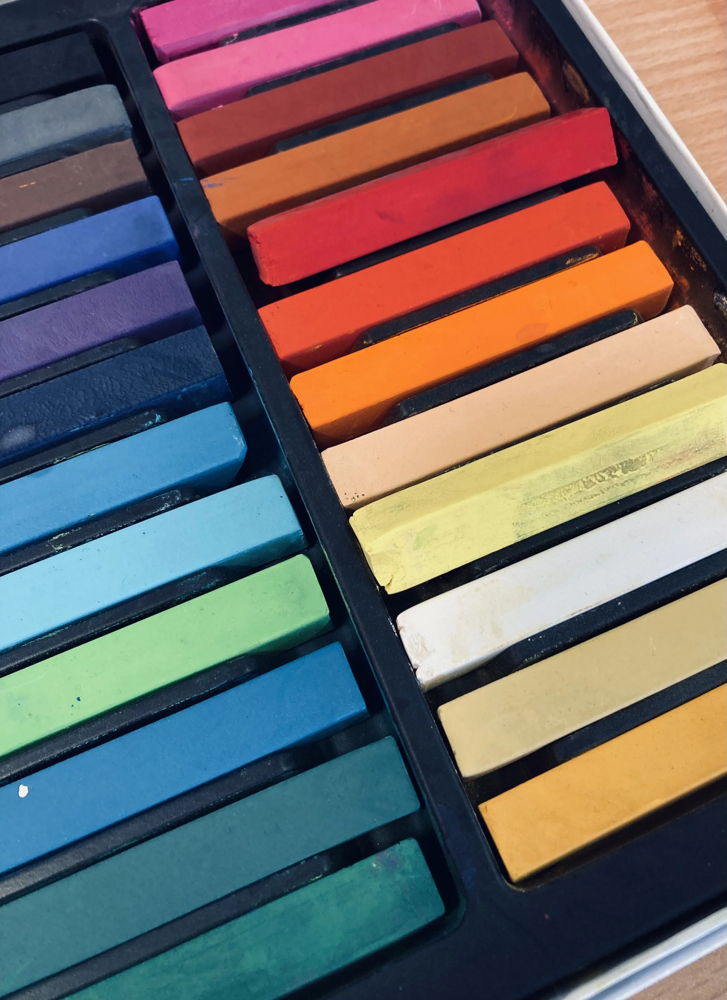 A set of colorful pastel chalksticks arranged in rows in a box, organized by color from blues and greens to reds and yellows.