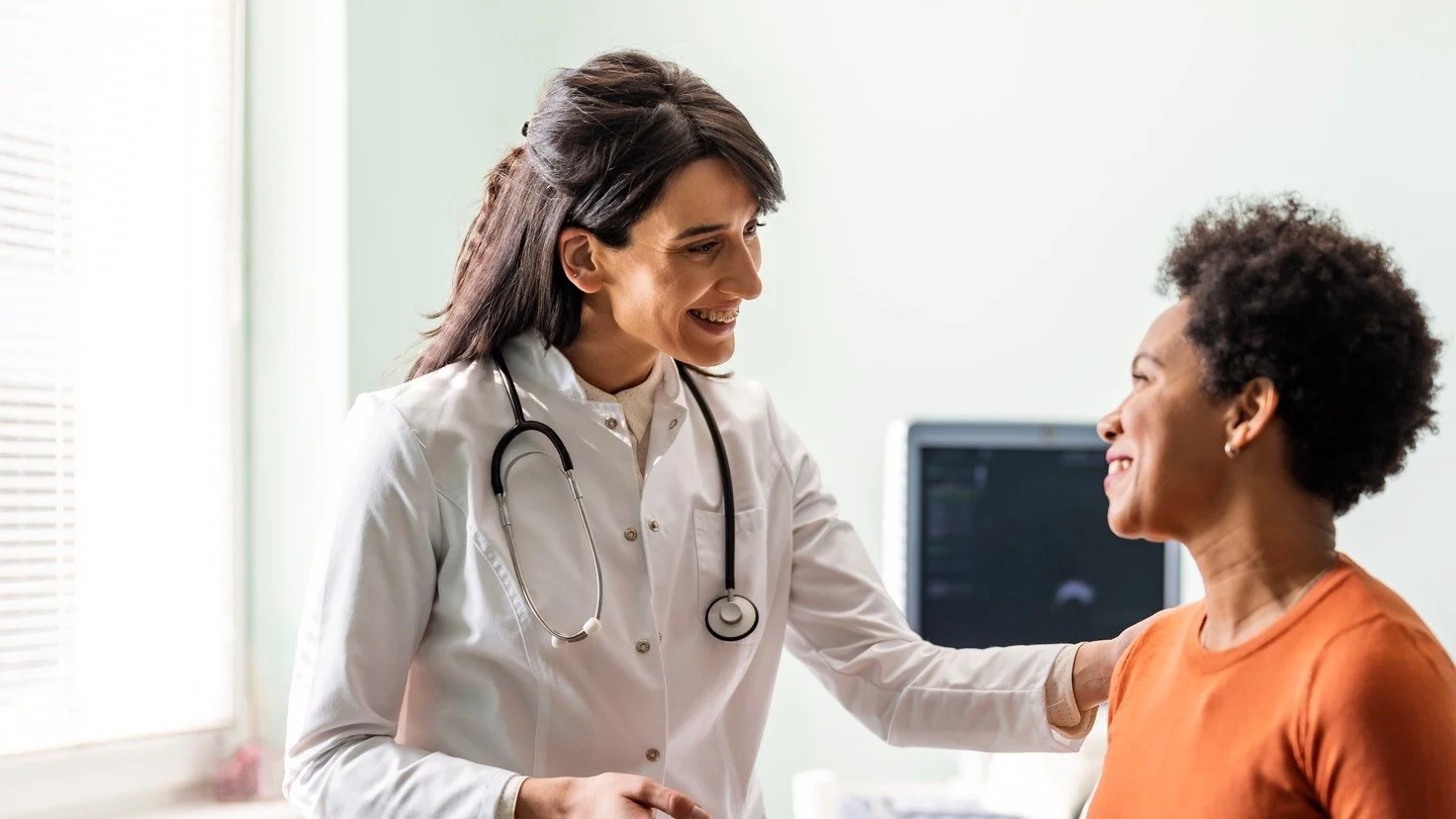A female doctor in a white coat with a stethoscope talking to a smiling female patient in an examination room.
