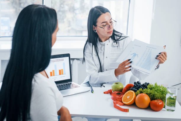 Two women in a bright office discussing diet plans, with various fruits on the table and a laptop displaying a bar chart.