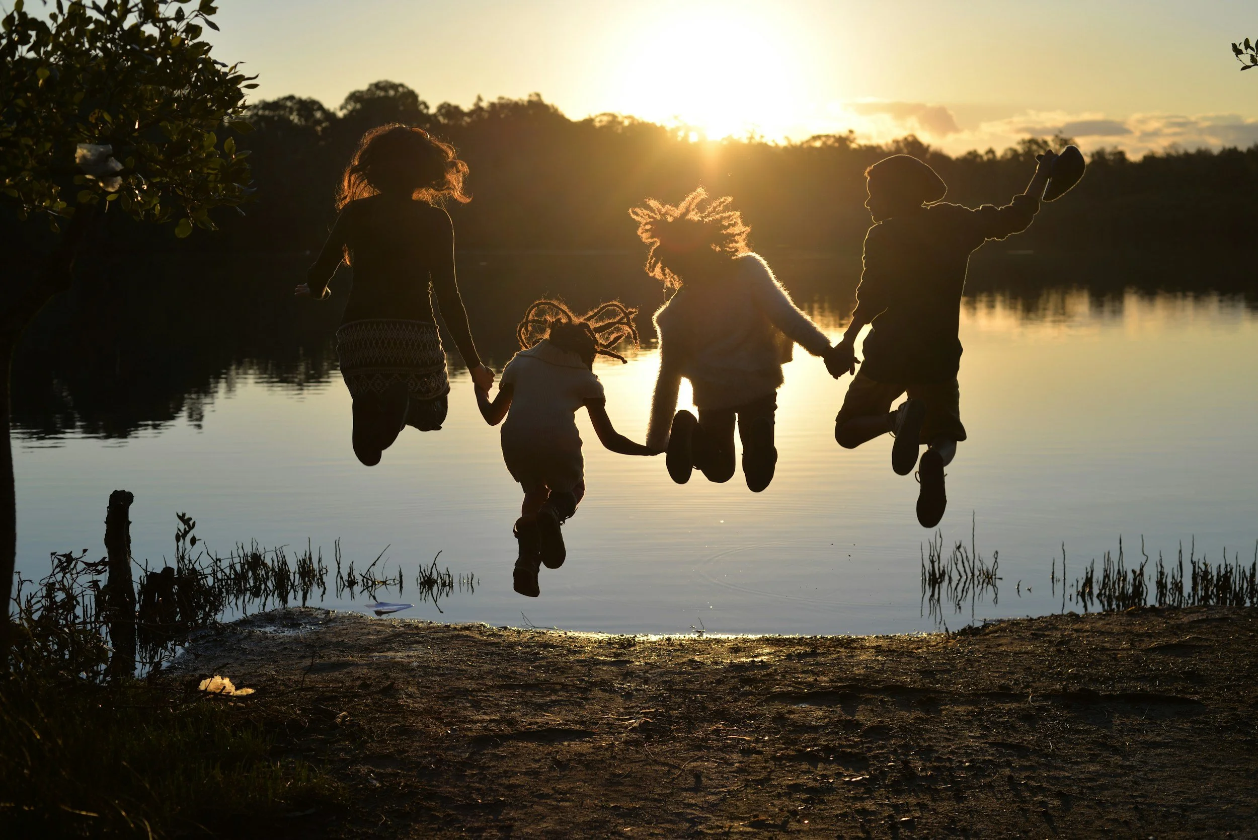 Silhouettes of five children jumping by a lake during sunset.