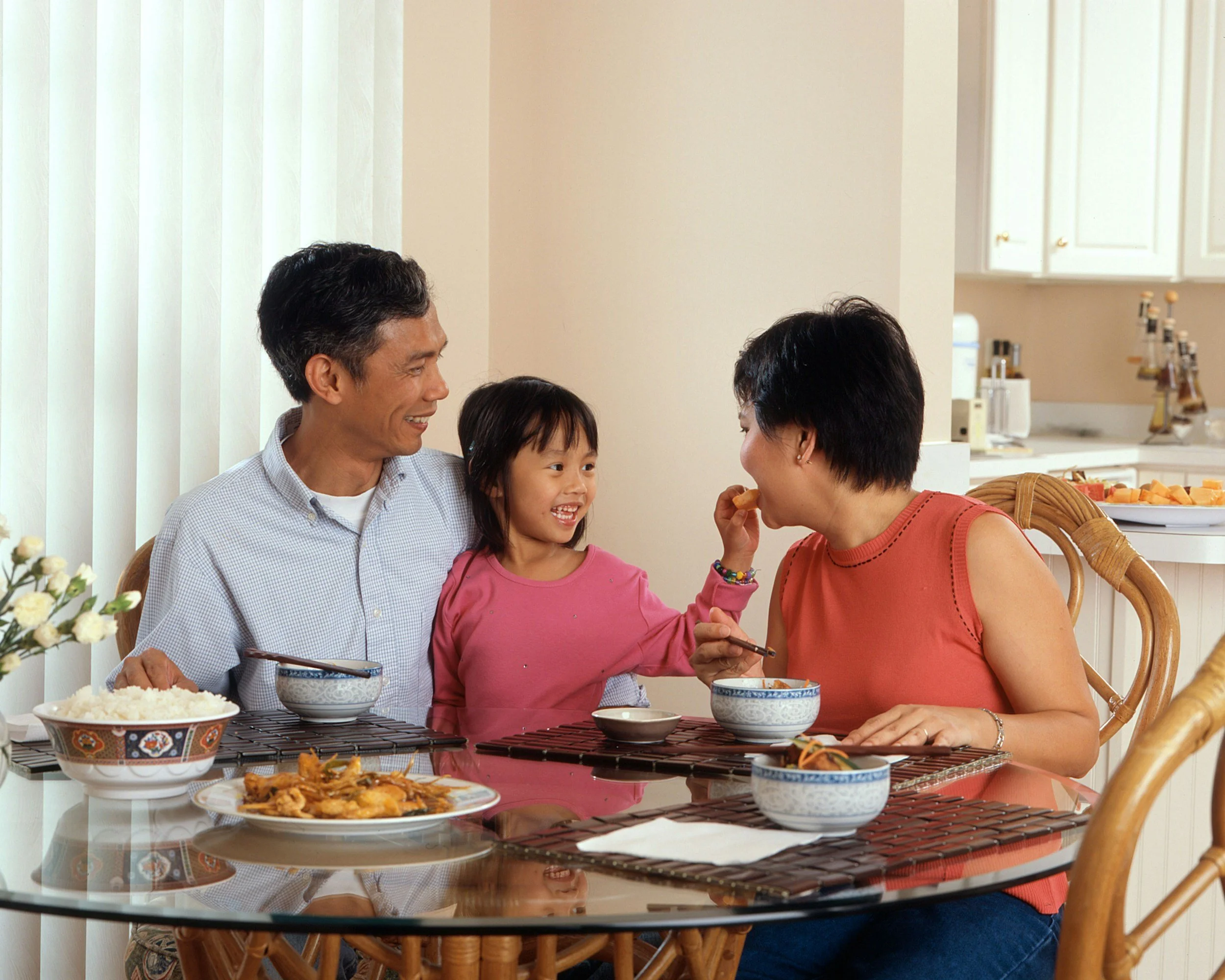 Family of three enjoying a meal together at the dining table, with a child playfully feeding the mother while the father smiles.