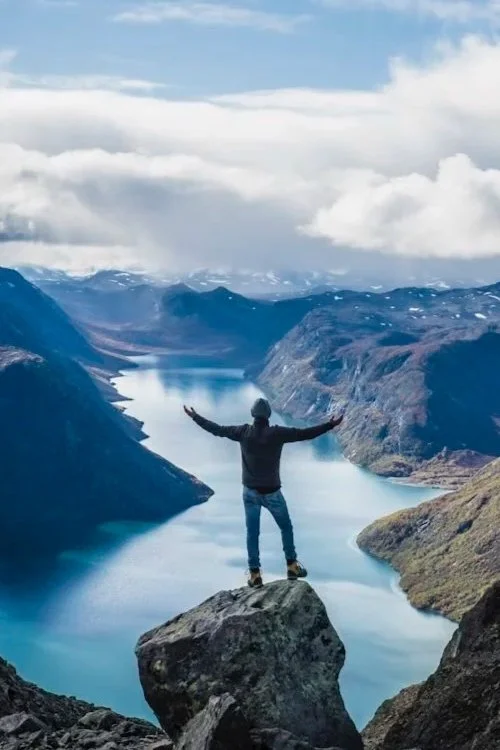 Persona de pie sobre una roca en un mirador natural, con brazos abiertos, rodeada de un paisaje montañoso y un lago en el valle.