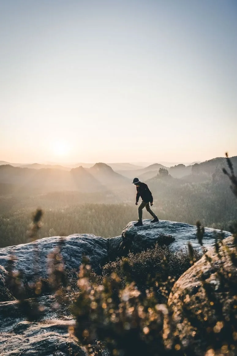 Persona caminando sobre rocas en un paisaje montañoso con amanecer o atardecer.