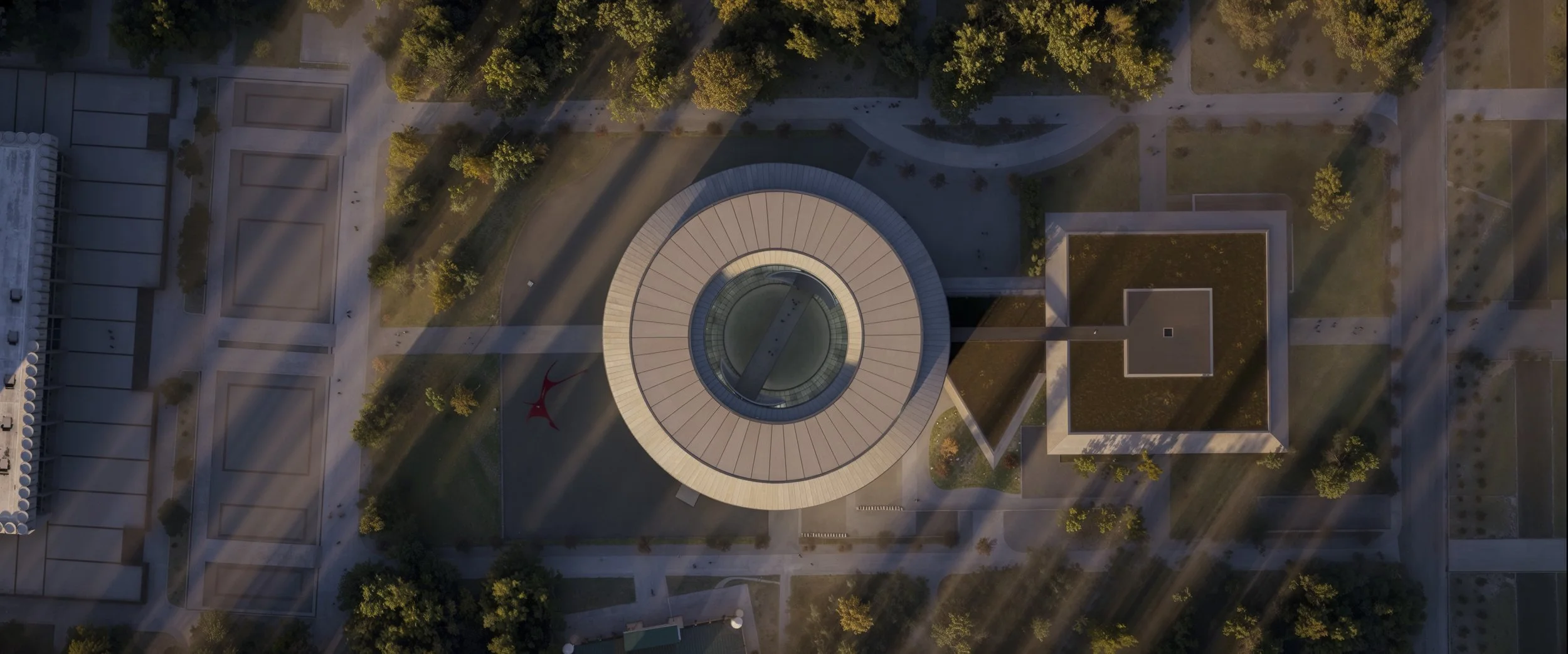 An aerial view of a modern circular building with a glass ceiling, surrounded by trees and pathways in a park or campus setting, during what appears to be late afternoon or early evening.