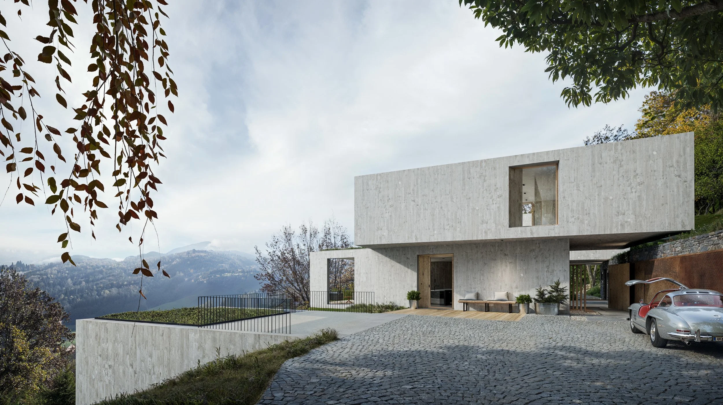 Modern concrete house with large windows, situated on a hill with a cobblestone driveway and scenic mountain view in the background. There is a parked car and outdoor seating area with potted plants.