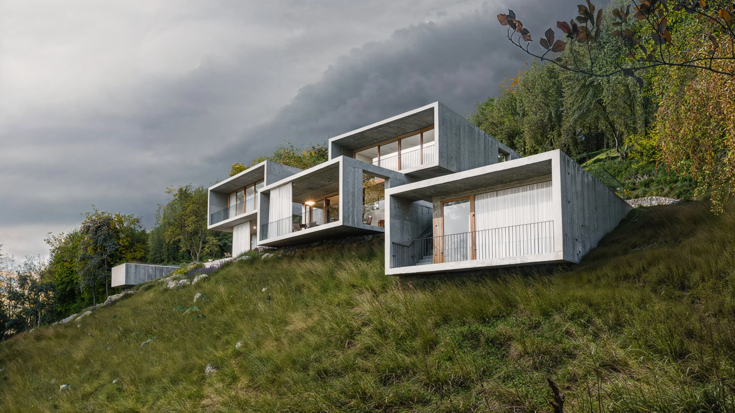 Modern concrete houses built on a hillside surrounded by trees and greenery, with a cloudy sky overhead.