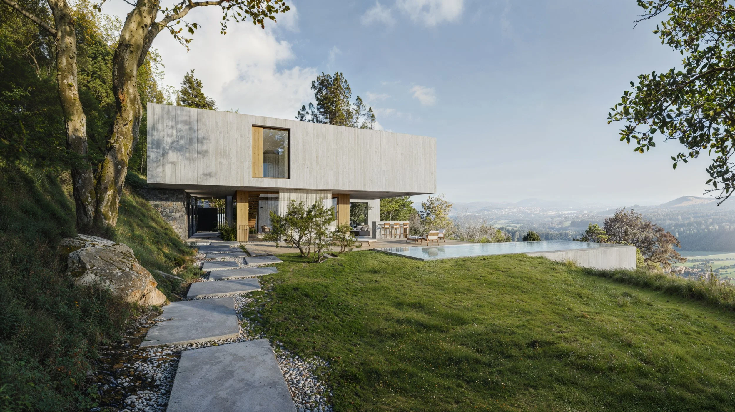 Modern house with concrete walls on a hillside, outdoor patio with seating and a pool, surrounded by trees and green grass, overlooking a valley with distant mountains.