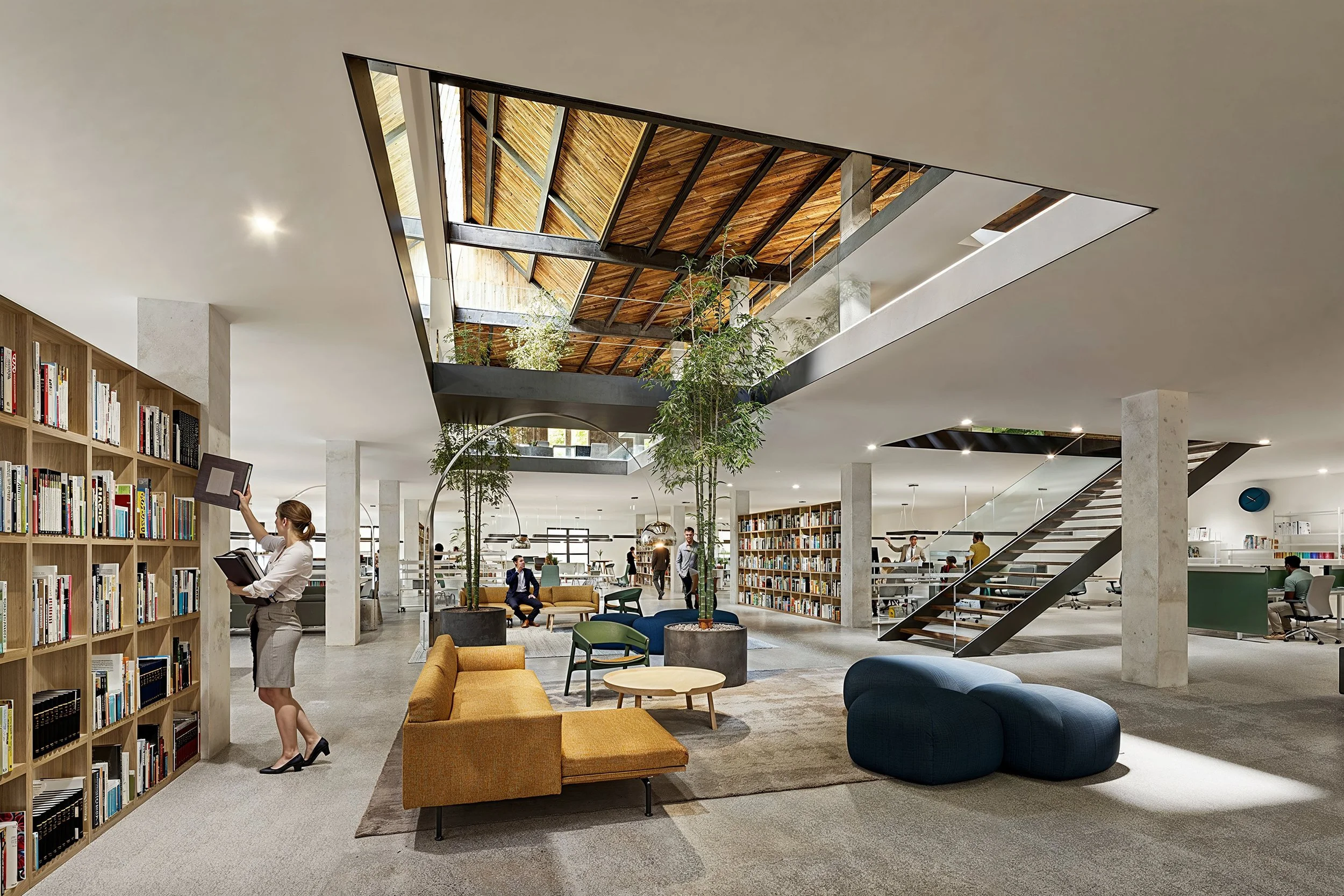 A modern library interior with a person browsing books on a wooden bookshelf, seating areas with colorful chairs and sofas, and an open space with a staircase and skylights.