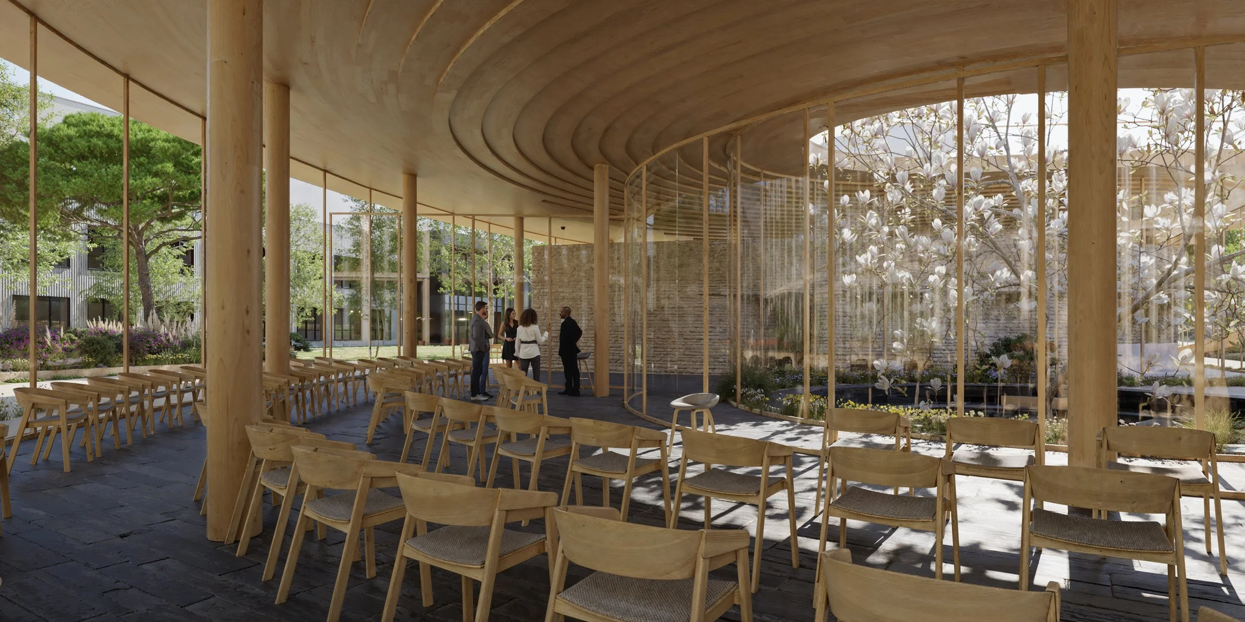 Interior of a modern building with wooden furniture and large glass windows, people gathered and talking in a bright, airy space with trees and plants visible outside.