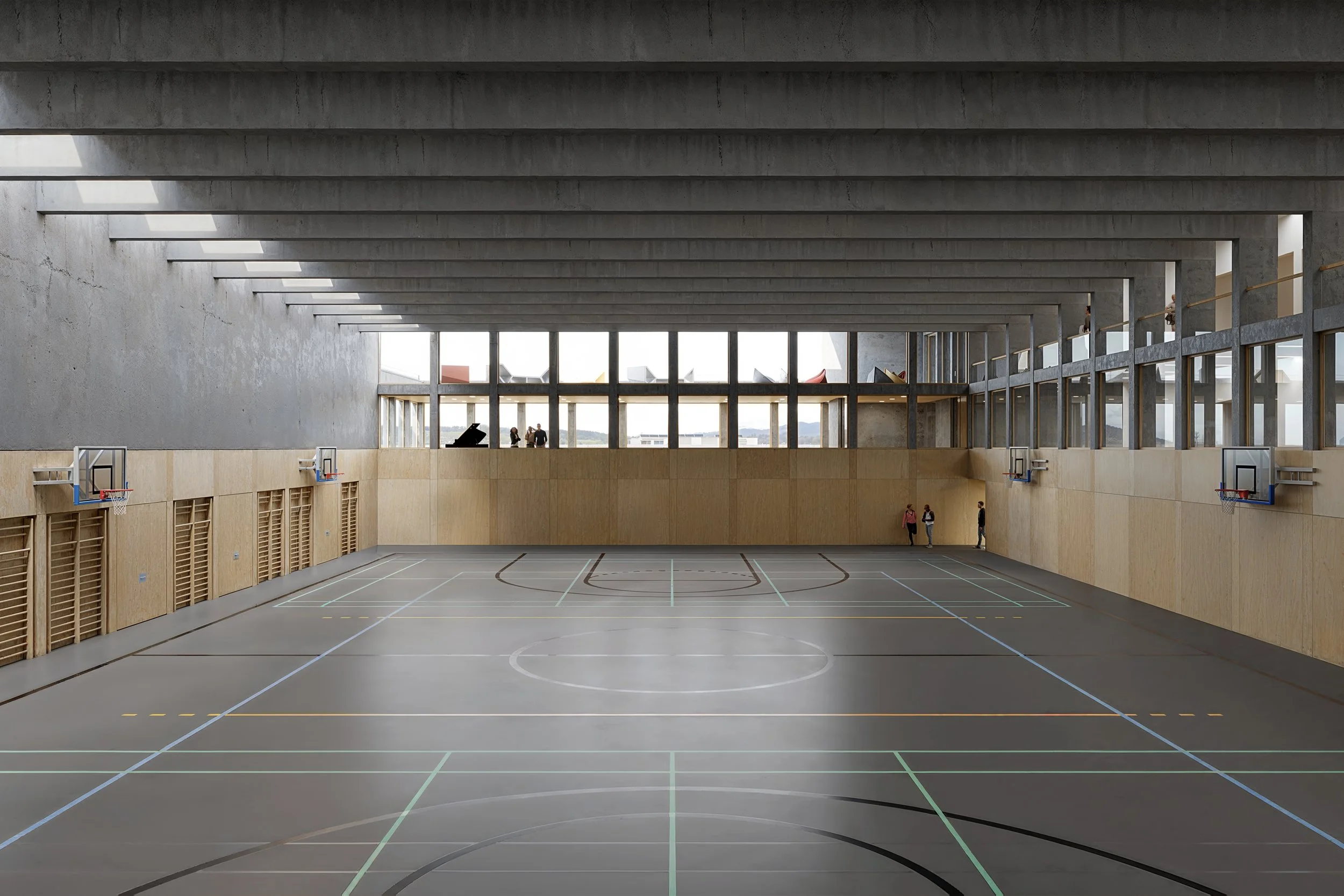Empty indoor basketball court with four basketball hoops, wooden walls, and large windows allowing natural light.