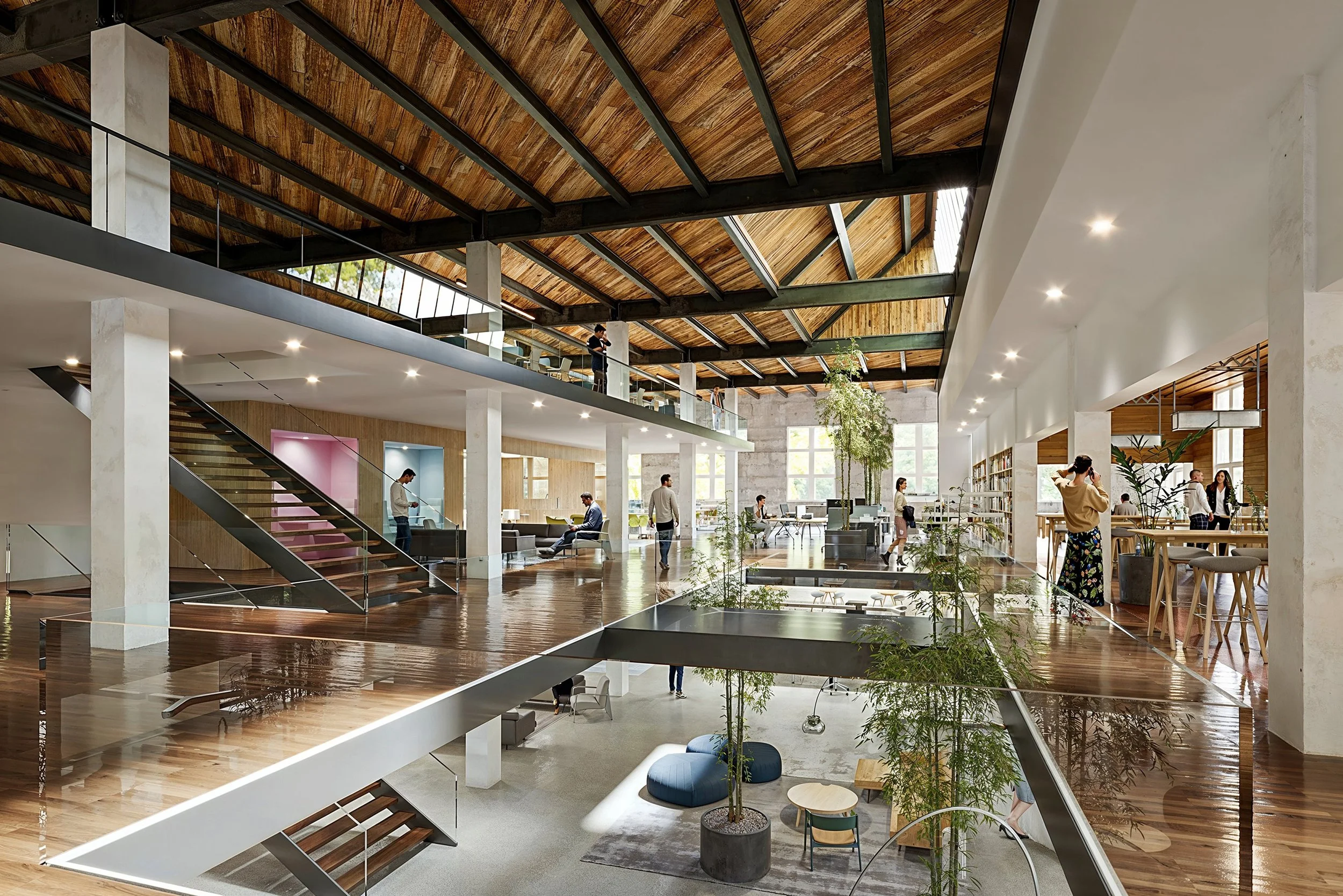 Interior view of a modern, open-concept office space with multiple levels, large windows, wooden flooring, and green plants. People are working and socializing in different areas, with a staircase connecting the floors.