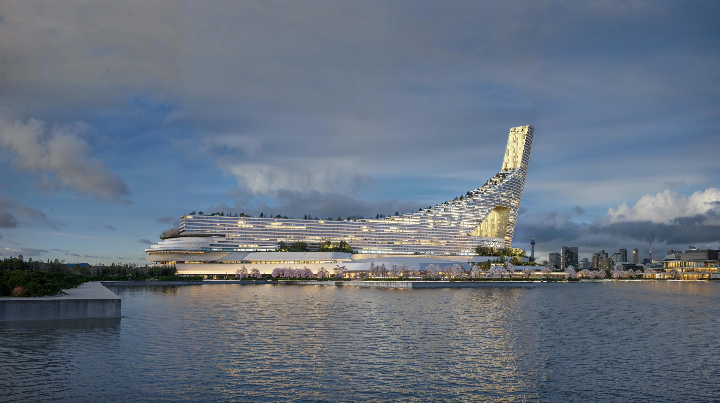 Modern building with a curved design and tower, situated next to a body of water with city skyline and cloudy sky in the background.