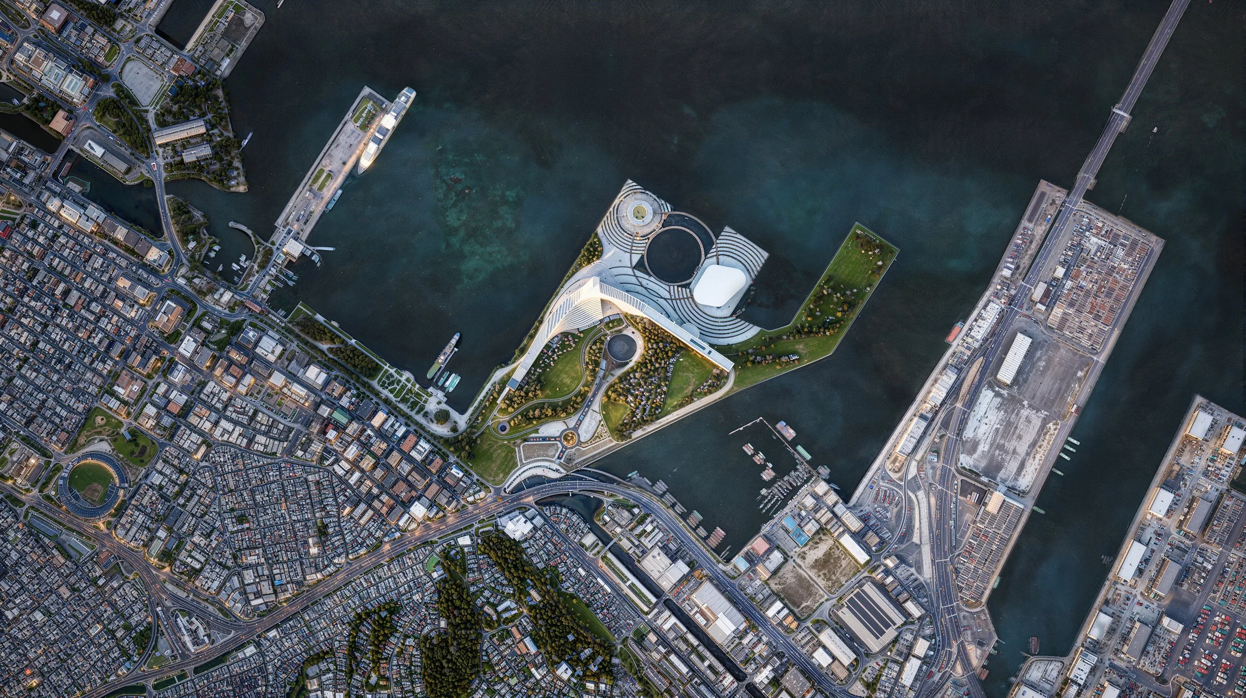 Aerial view of city with water, bridges, and modern buildings, including a prominent uniquely shaped building with surrounding greenery.