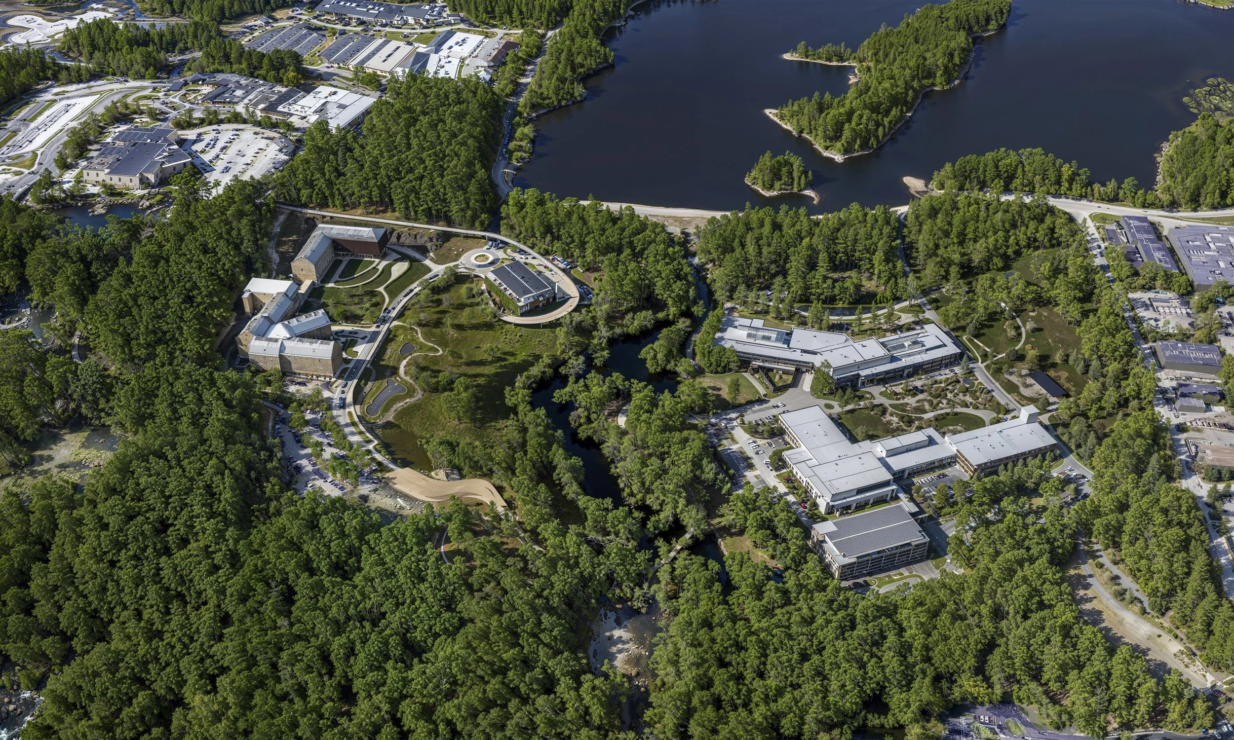 An aerial view of a campus with multiple buildings surrounded by dense green trees, water bodies, and parking lots.