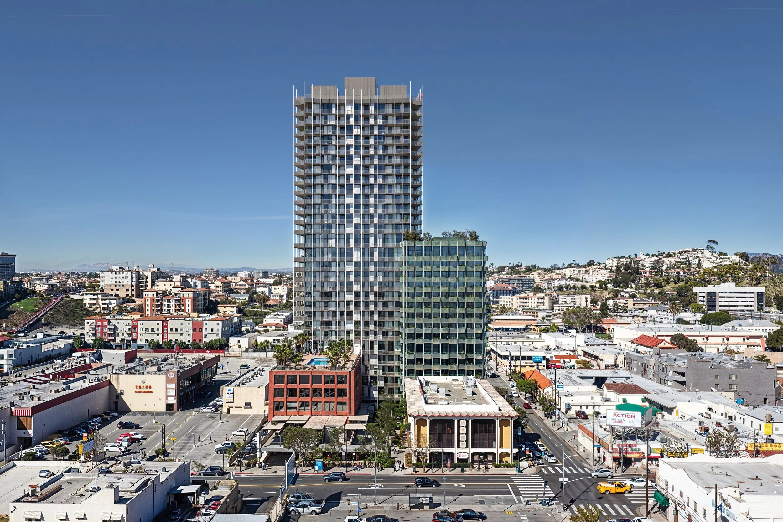 A cityscape featuring a tall modern high-rise building in the center, surrounded by various smaller buildings, streets, and parked cars on a sunny day with a clear blue sky.