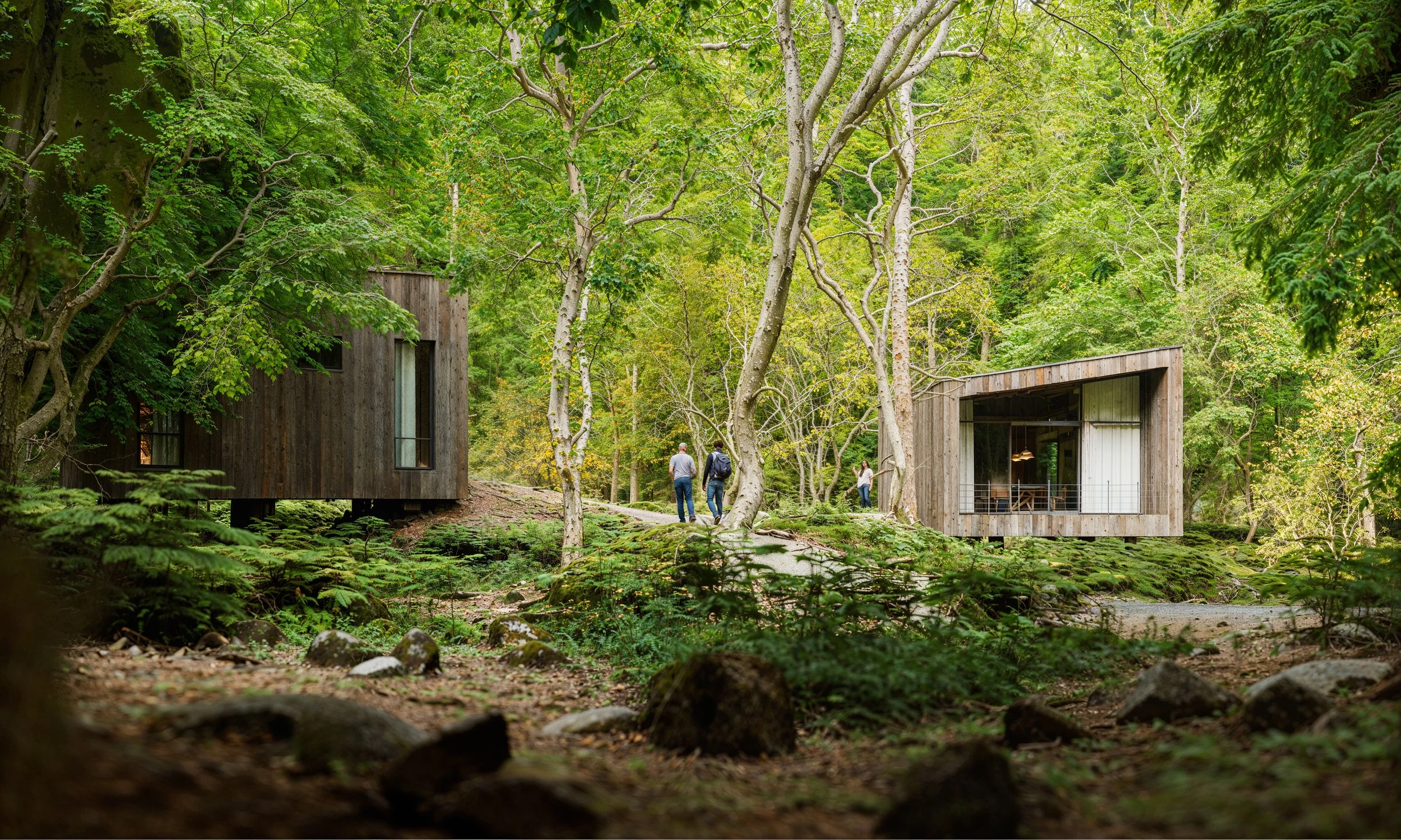 Two modern houses with wood exteriors are situated among green trees in a forest. Three people are walking on a path between the houses.