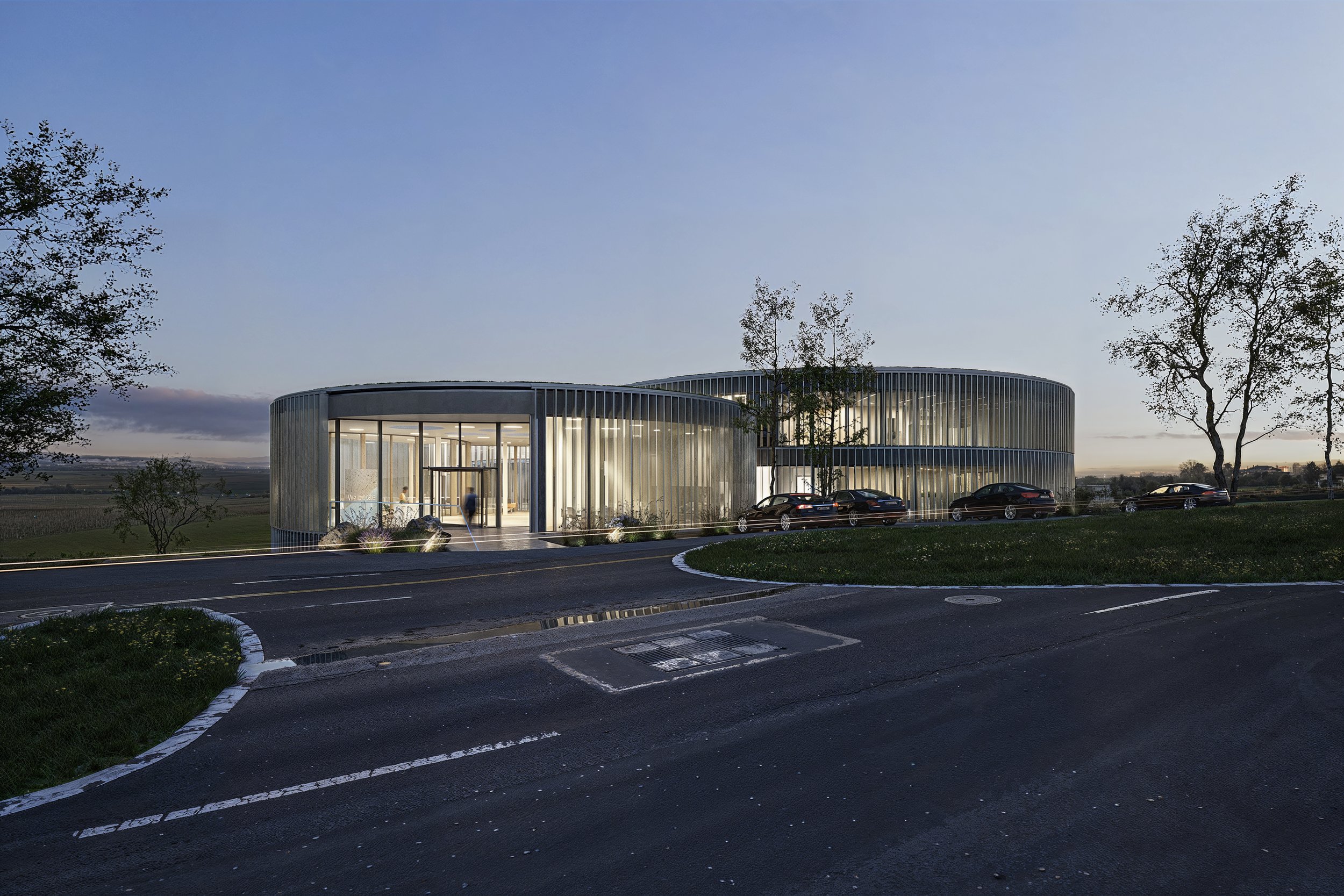 Modern circular office building with large glass windows and vertical metal slats, illuminated from inside, with parked cars and trees in the surrounding area at dusk.