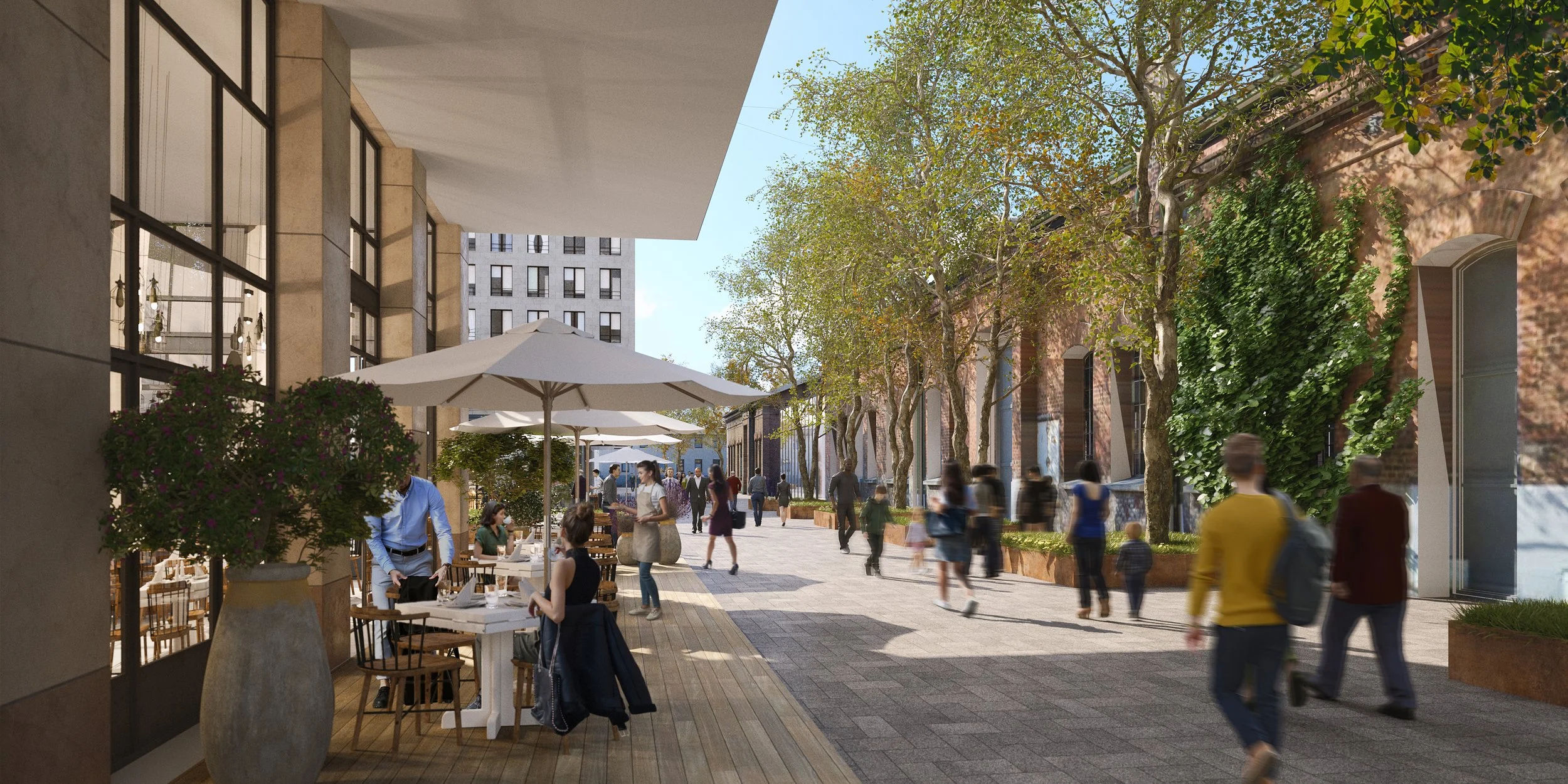 Urban outdoor shopping and dining area with people walking, sitting, and chatting, surrounded by trees, buildings, and outdoor tables with umbrellas, in daylight.